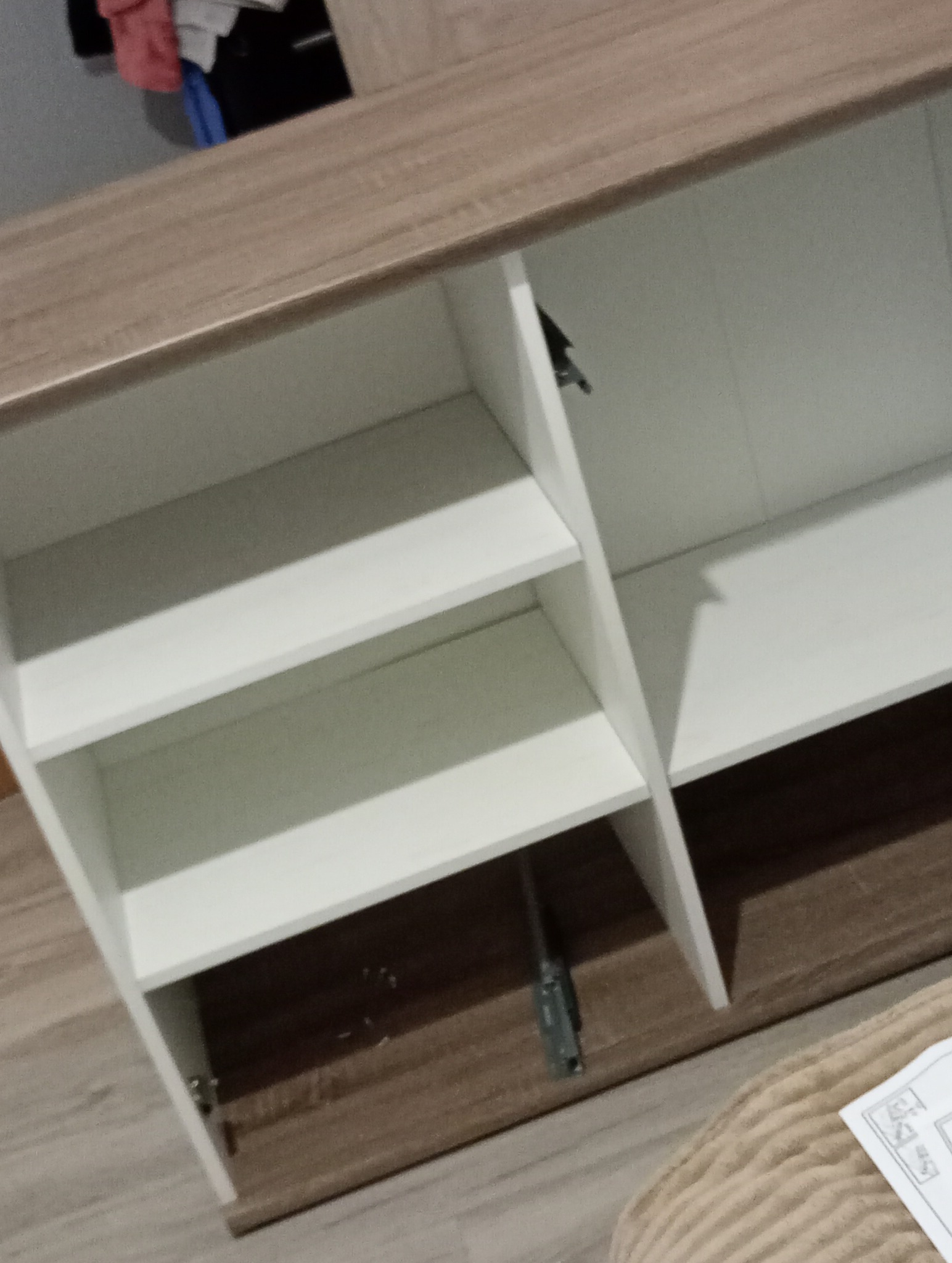 Wnętrze szafki z półkami, białe fronty i jasnobrązowy korpus, widoczne okucia i zawiasy, częściowo otwarte drzwiczki.