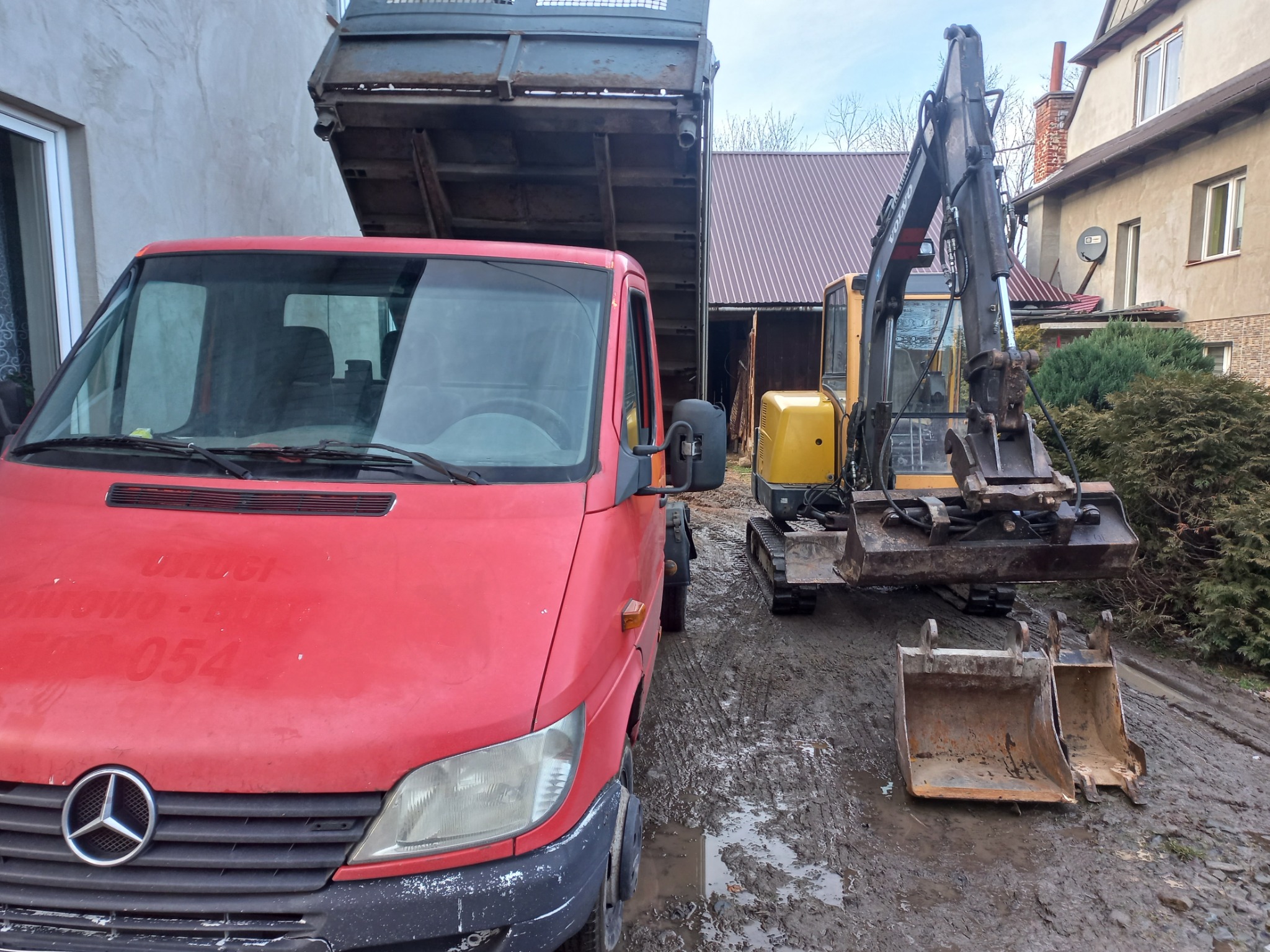Czerwony samochód ciężarowy Mercedes-Benz z podniesioną skrzynią ładunkową, zaparkowany obok żółtej koparki na gąsienicach i dwóch łyżek koparki na błotnistym terenie.