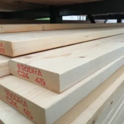 Stos jasnej, świeżo przetartej tarcicy budowlanej z czerwonymi oznaczeniami jakości C24 45 na końcach, ułożonej na półce w magazynie.