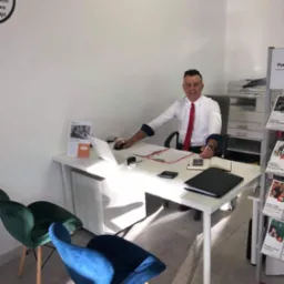 Uśmiechnięty doradca w białej koszuli i czerwonym krawacie siedzi za biurkiem w jasnym biurze, przed nim laptop, dokumenty, kalkulator i pieczątka, w tle drukarka i ulotki reklamowe.