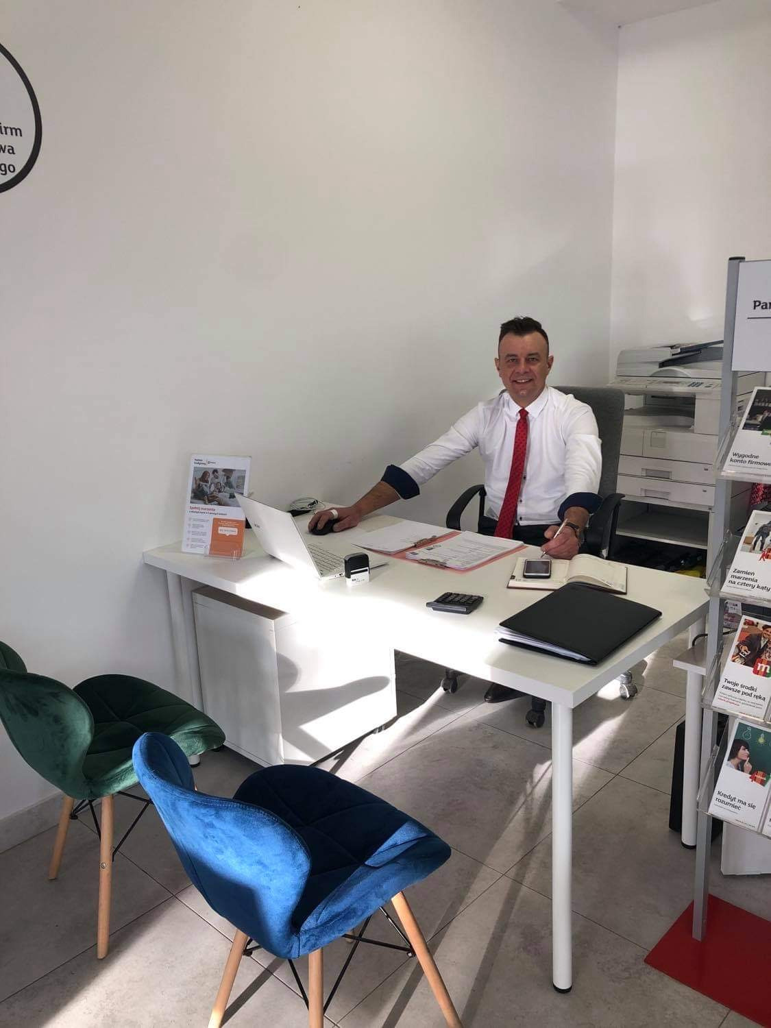 Uśmiechnięty doradca w białej koszuli i czerwonym krawacie siedzi za biurkiem w jasnym biurze, przed nim laptop, dokumenty, kalkulator i pieczątka, w tle drukarka i ulotki reklamowe.