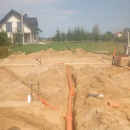 Wykonane wykopy pod fundamenty domu jednorodzinnego z widocznymi rurami kanalizacyjnymi w wykopach, na tle nowy dom w stanie surowym otwartym.
