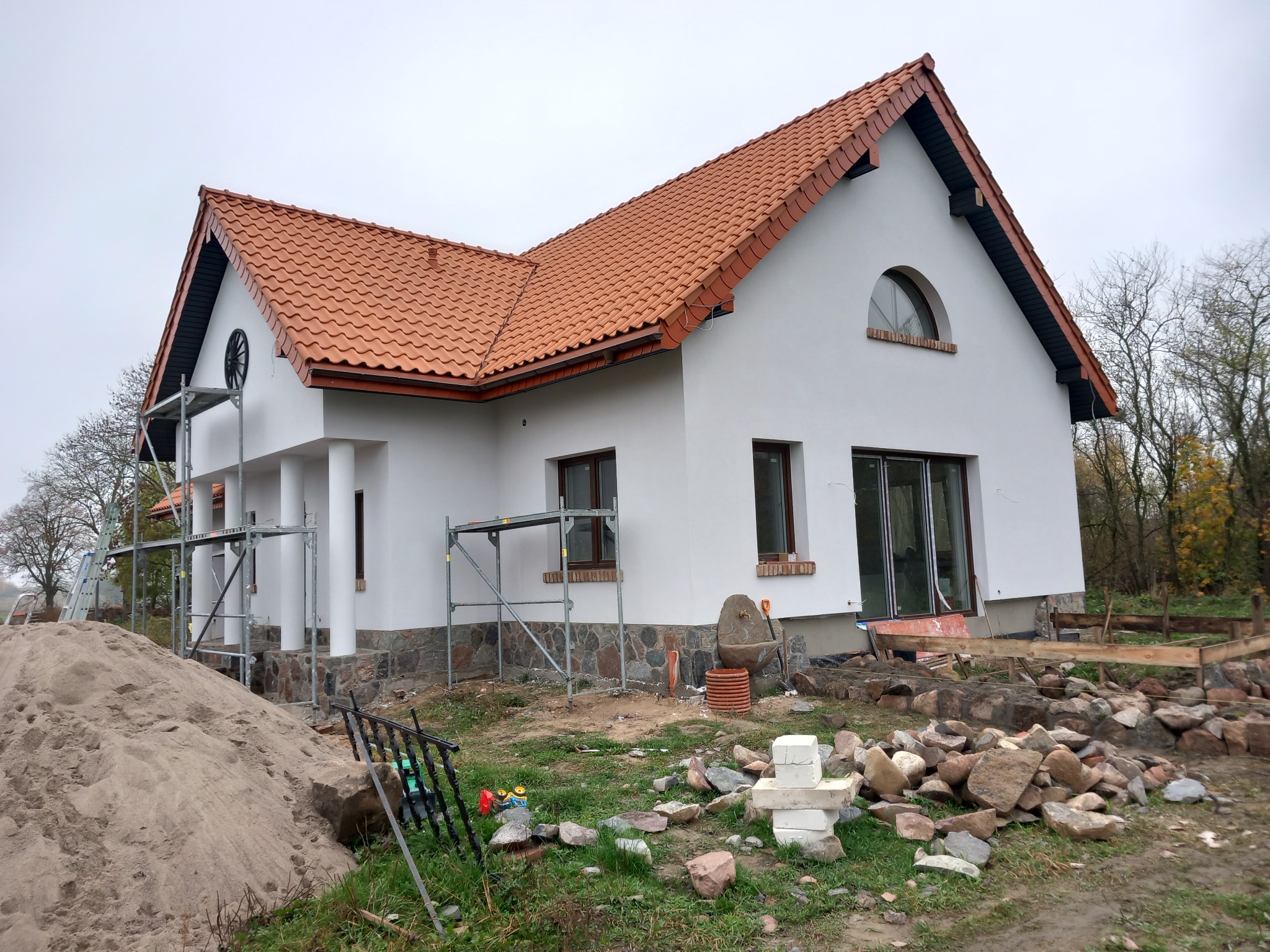 Nowoczesny dom jednorodzinny z czerwoną dachówką w trakcie wykończenia elewacji, z widocznymi rusztowaniami, stertą piasku i kamieniami na trawniku.