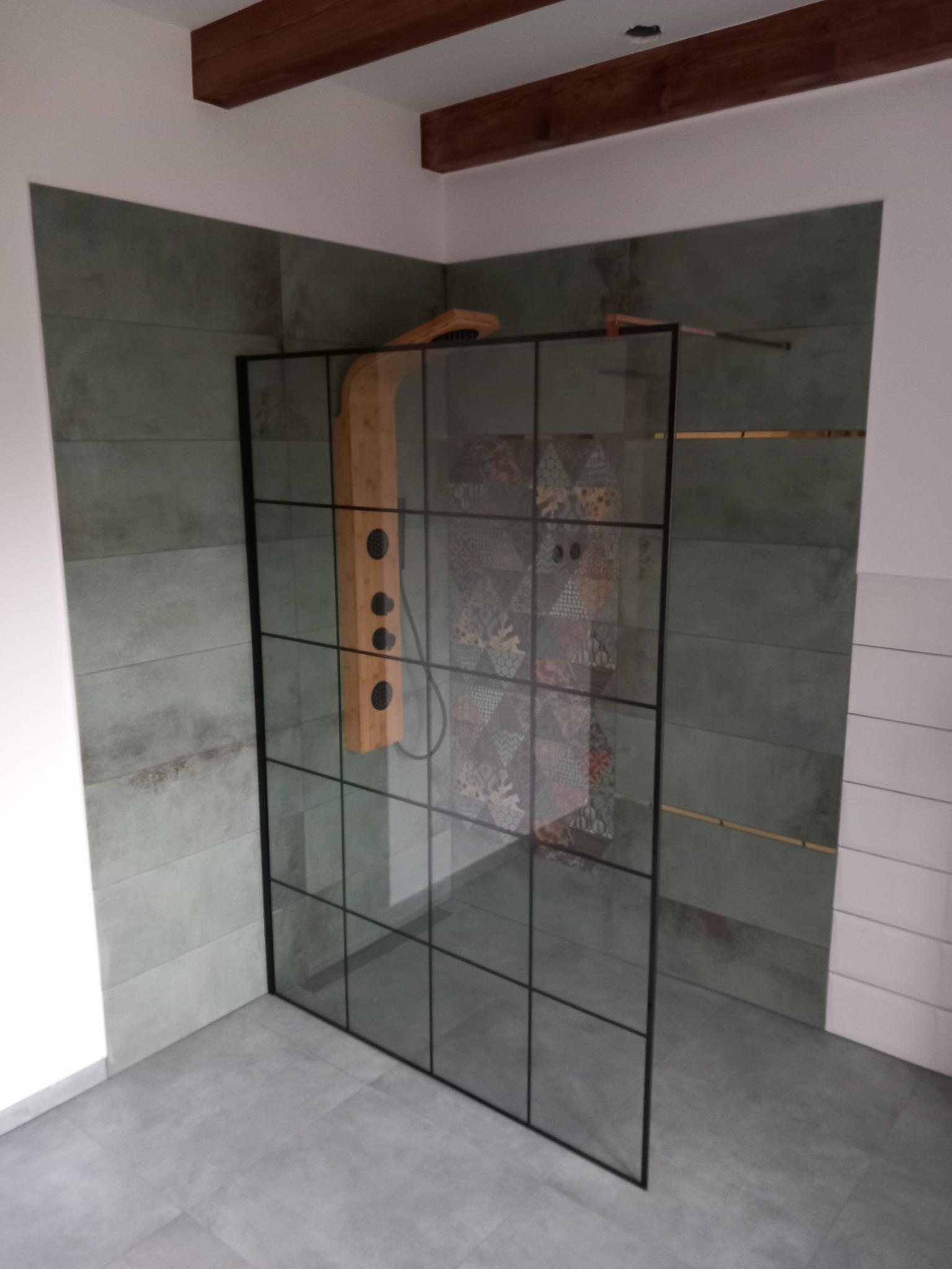 Nowoczesna kabina prysznicowa typu walk-in z czarnymi profilami, drewnianą deszczownicą i panelami ściennymi w odcieniach szarości i zieleni, wzorzyste płytki widoczne przez szybę, sufit...