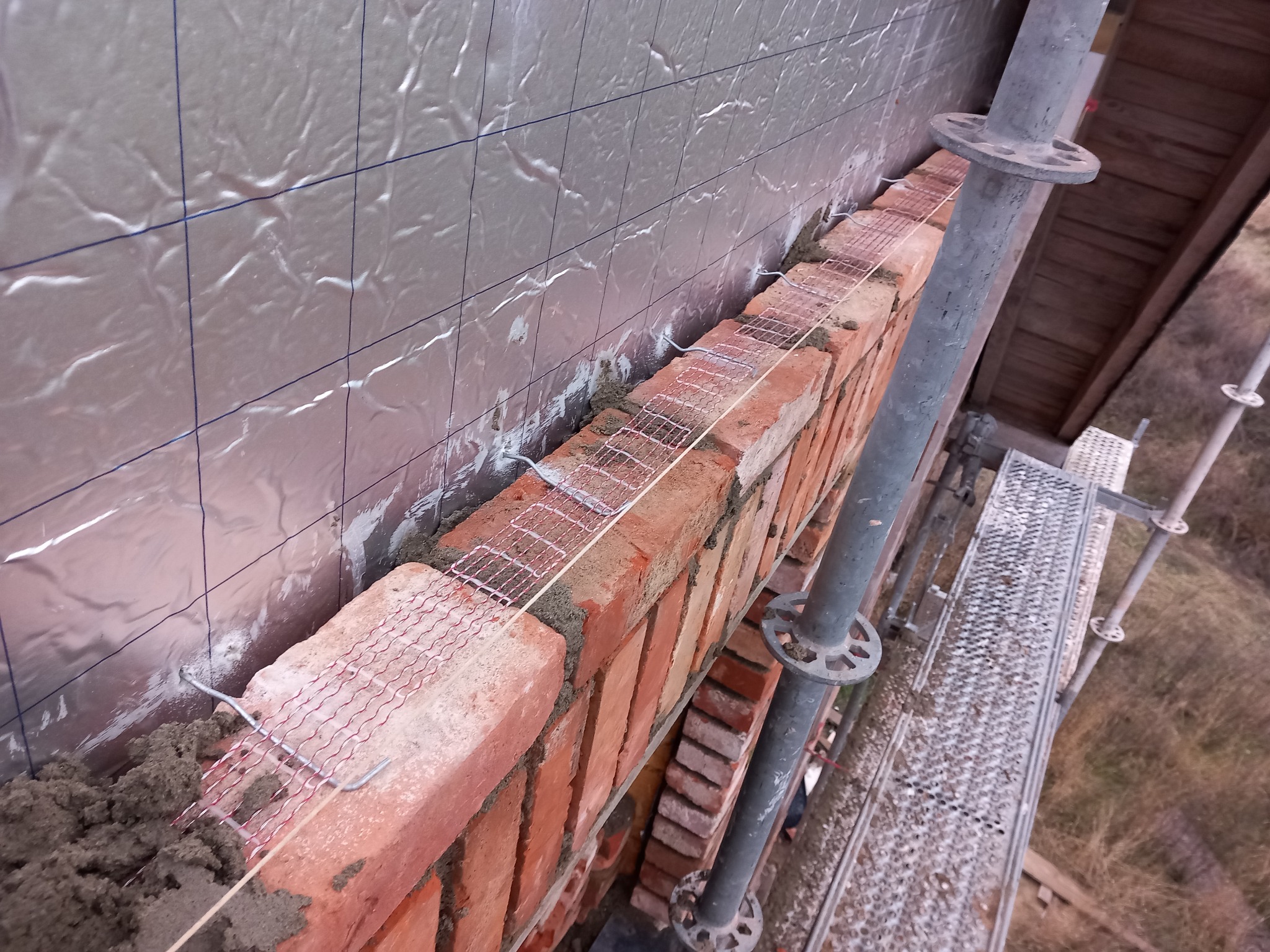 Wznoszenie muru z czerwonej cegły z metalową siatką wzmacniającą na tle srebrnej folii izolacyjnej z niebieską siatką, widok z rusztowania.