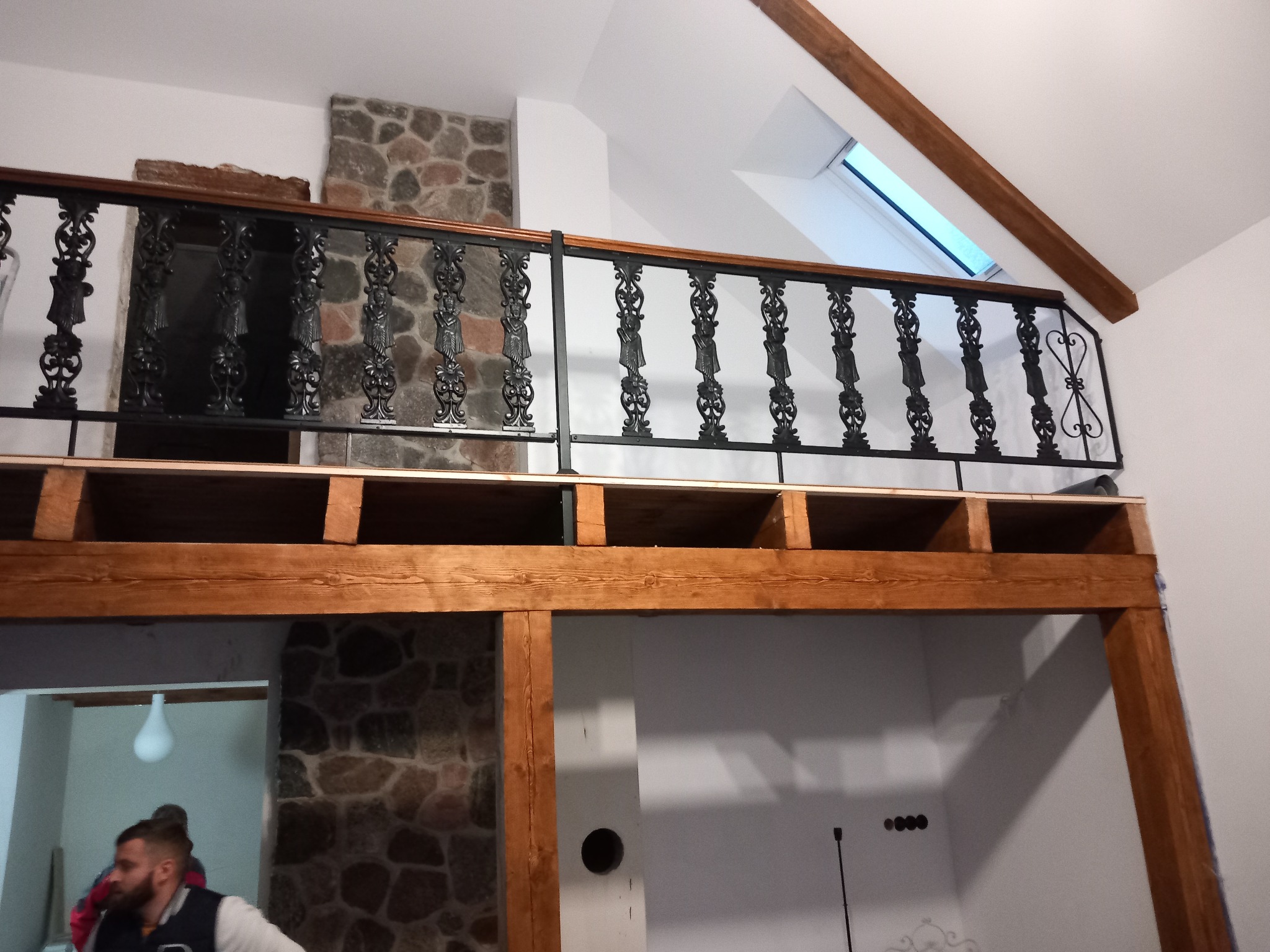 Wnętrze domu w trakcie tworzenia antresoli z drewnianą konstrukcją i ozdobną, kutą balustradą, widoczne fragmenty kamiennej ściany i okno dachowe.
