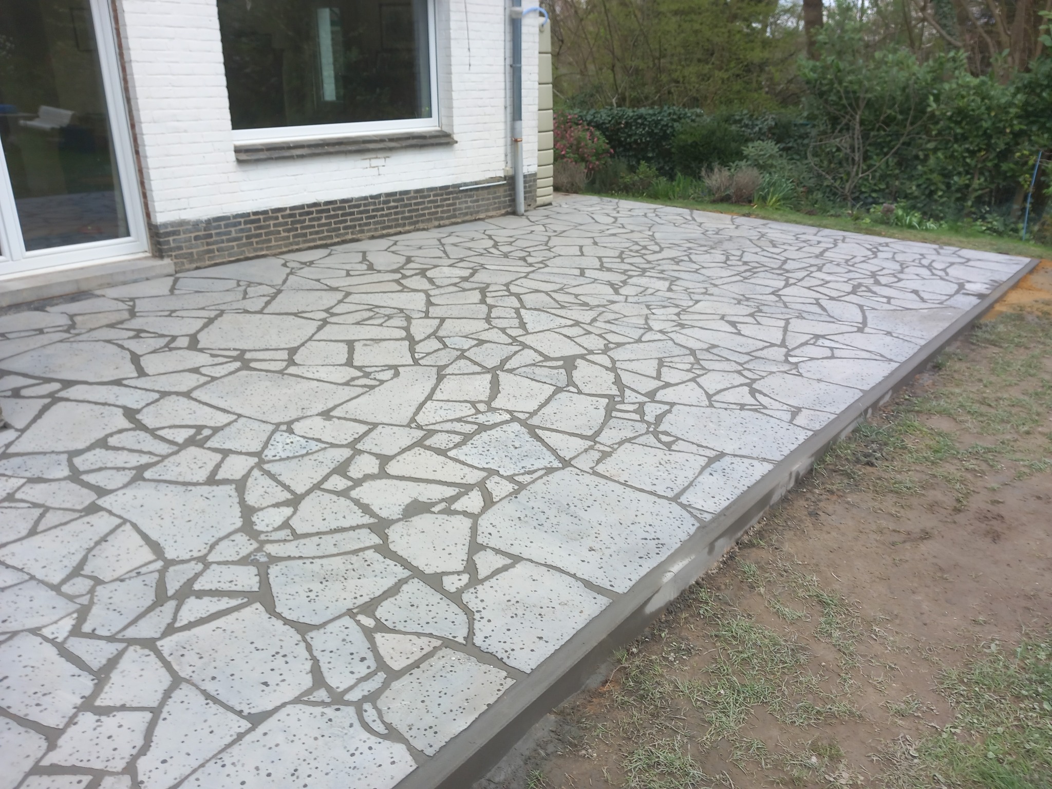 Wykończony taras z nieregularnych płyt kamiennych w odcieniach szarości, przylegający do białego budynku z oknem i drzwiami balkonowymi, otoczony zielenią.