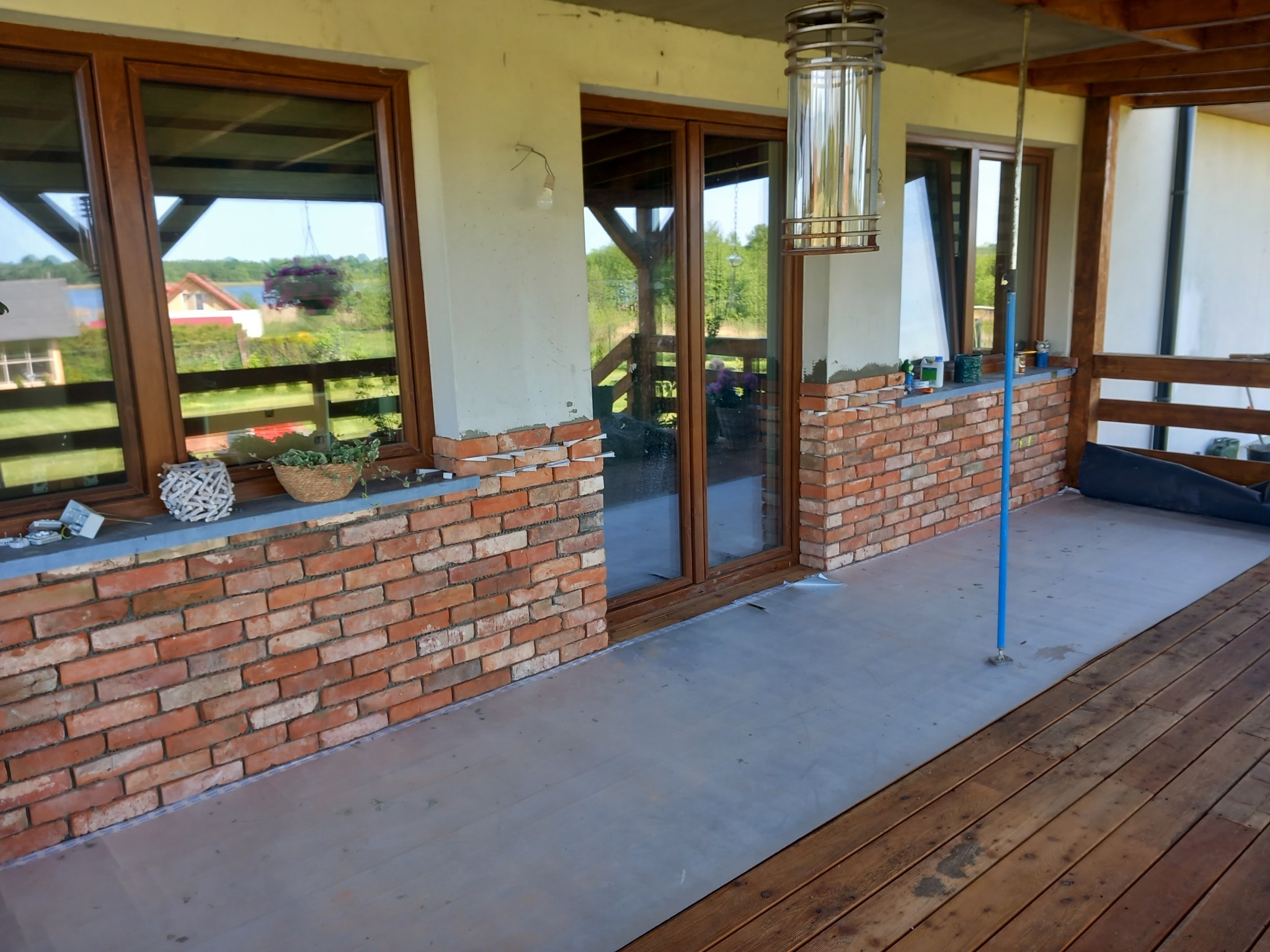 Wykończony taras z ceglaną elewacją, okna z brązowymi ramami, zabezpieczona folią podłoga, widoczna lampa wisząca i elementy dekoracyjne na parapecie.