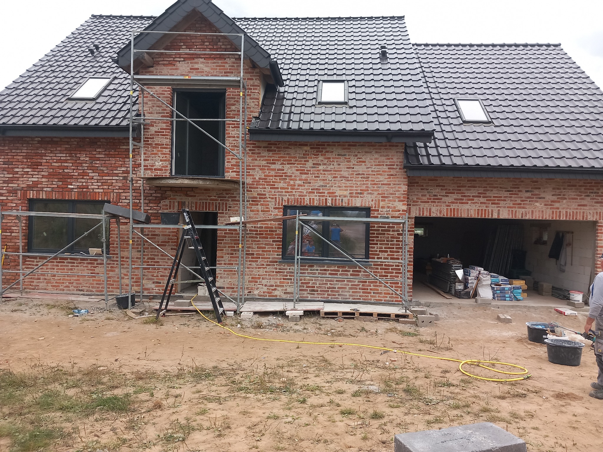 Surowy stan domu z czerwonej cegły w Ełku, z czarną dachówką, w trakcie budowy, z widocznym rusztowaniem i materiałami budowlanymi.