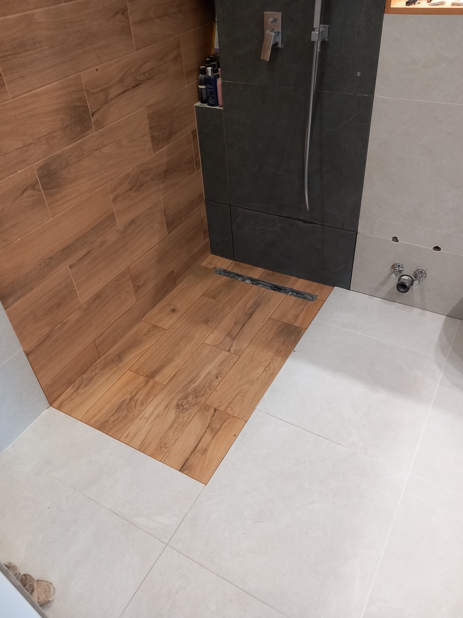 Fragment łazienki z drewnopodobnymi płytkami na ścianie i podłodze w strefie prysznica, kontrastującymi z szarymi płytkami na ścianie i jasnymi płytkami na podłodze, widoczny odpływ liniowy...