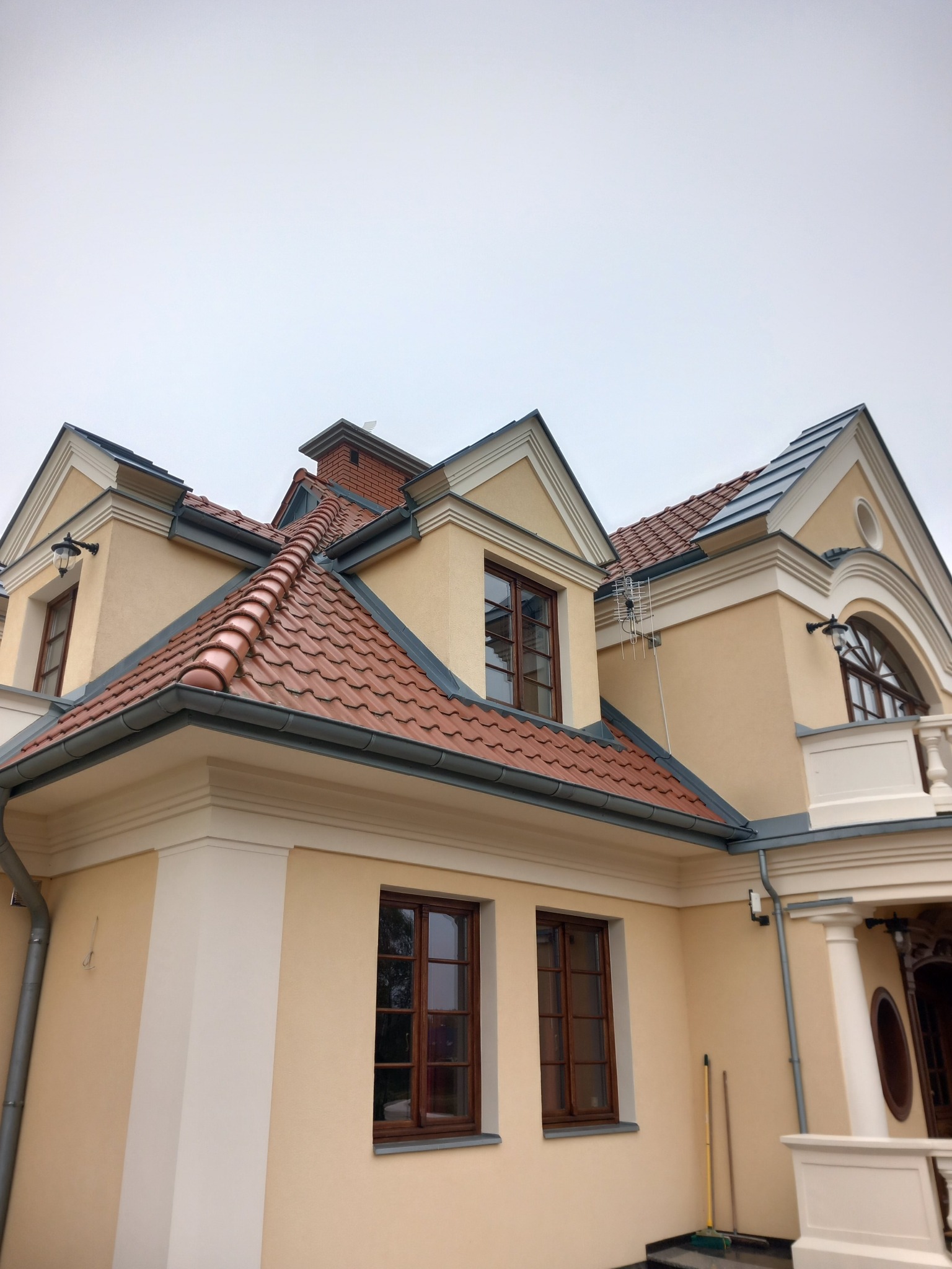Elewacja domu w odcieniach beżu z brązową dachówką i drewnianą stolarką okienną, widok z dołu na fragment budynku z widoczną anteną satelitarną.