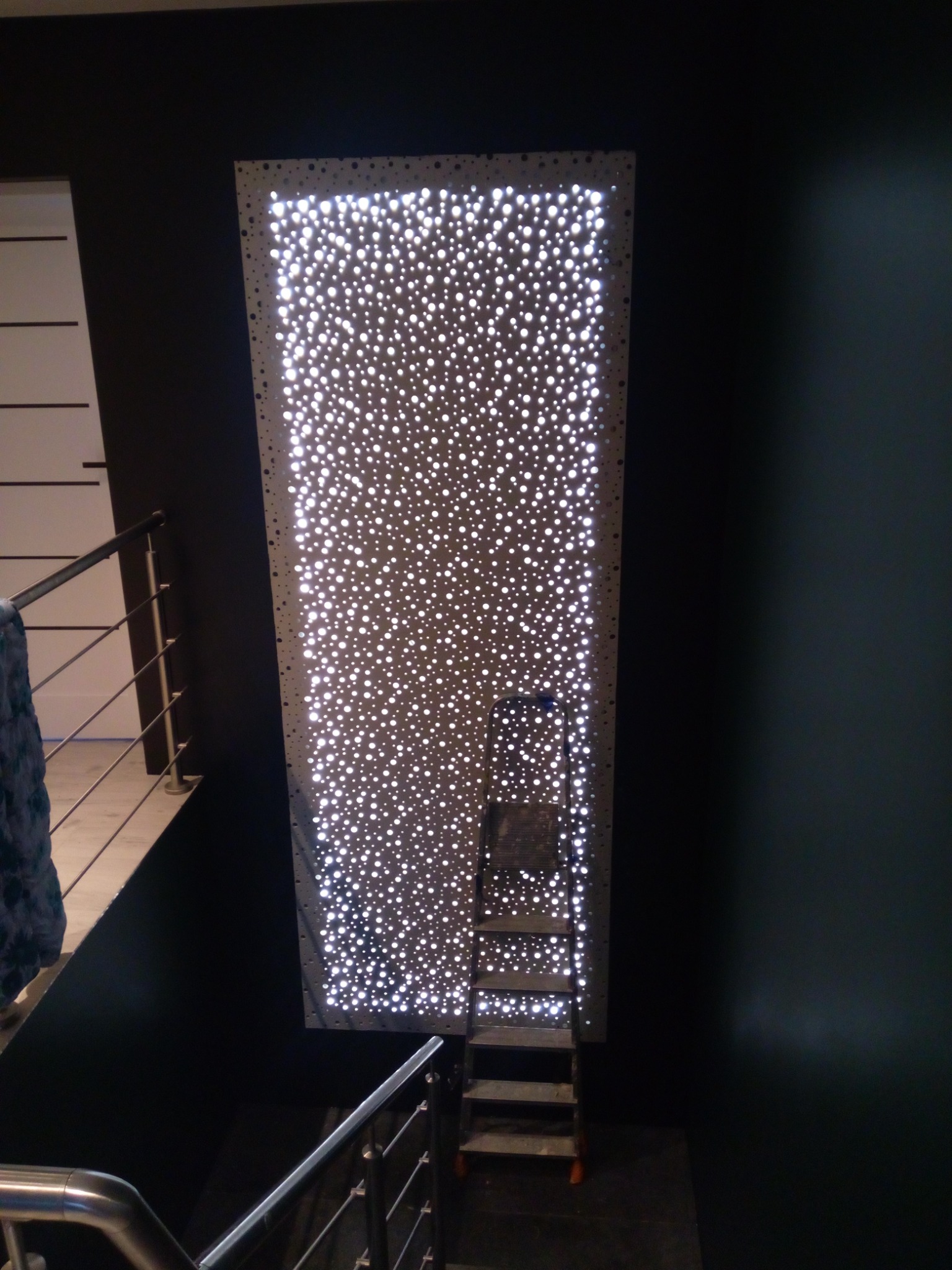 Podświetlana, perforowana płyta dekoracyjna na ścianie, drabina aluminiowa oparta o płytę, widok z góry ze schodów.