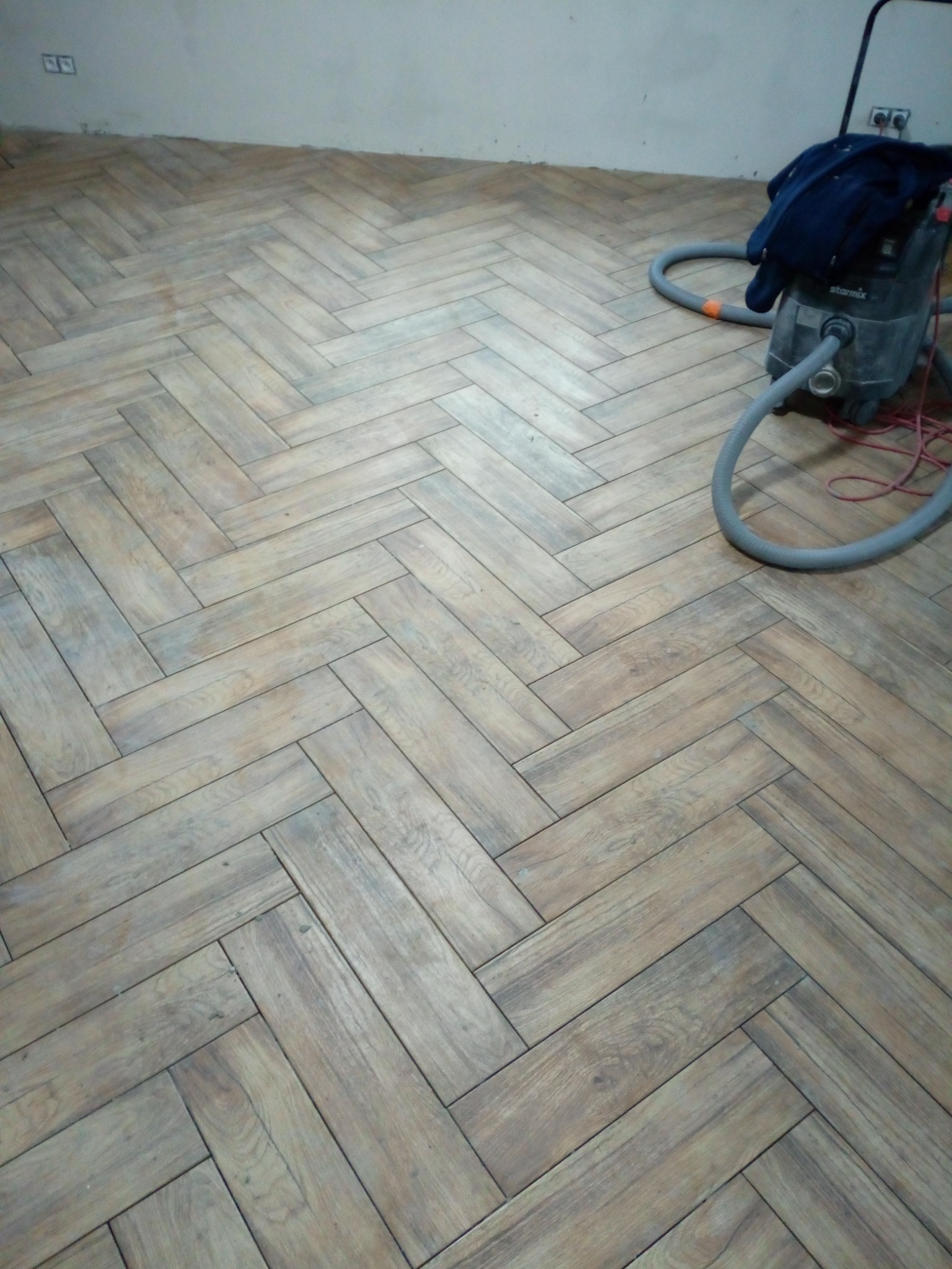 Ułożona podłoga z paneli w jodełkę, widoczny odkurzacz przemysłowy z wężem i kablem na podłodze, fragment ściany w tle.