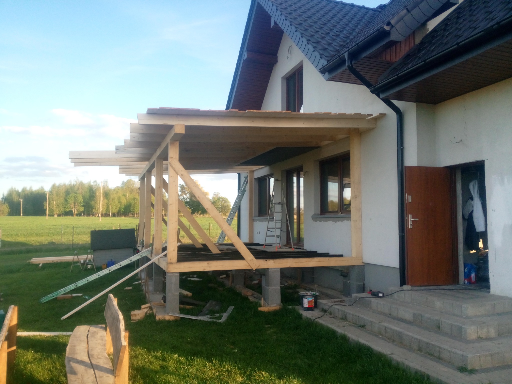 Budowa drewnianego zadaszenia tarasu przy domu jednorodzinnym, widoczne słupy i belki konstrukcyjne, częściowo ułożona podłoga, drabina oparta o ścianę, trawnik i drzewa w tle.