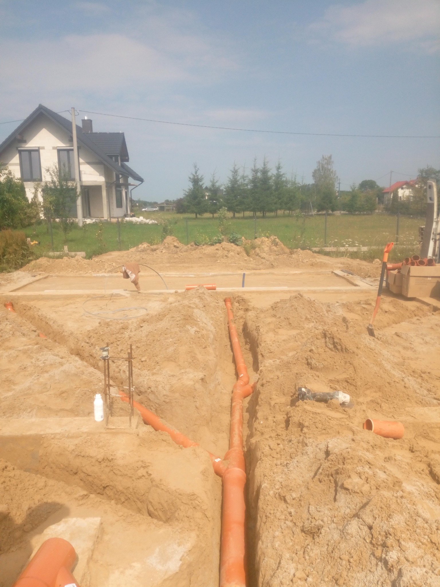 Wykonane wykopy pod fundamenty domu jednorodzinnego z widocznymi rurami kanalizacyjnymi w wykopach, na tle nowy dom w stanie surowym otwartym.