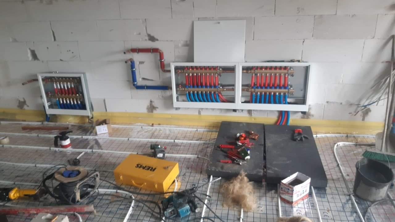 Montaż rozdzielaczy ogrzewania podłogowego, widoczne narzędzia, żółty kufer REMS, oraz instalacja rur czerwonych i niebieskich na ścianie z betonu komórkowego.