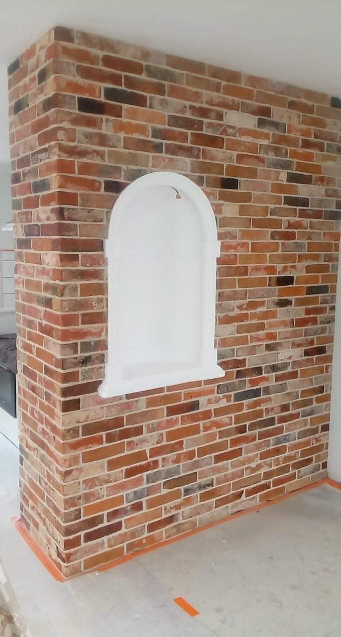 Fragment ściany z czerwonej cegły z wnęką o białym, łukowatym obramowaniu, zabezpieczona pomarańczową taśmą malarską przy podłodze.