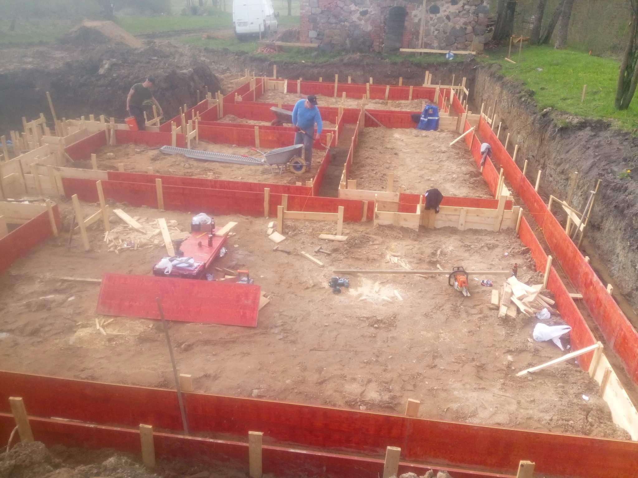 Budowa fundamentów domu jednorodzinnego z użyciem szalunków systemowych z czerwonymi panelami, widoczni pracownicy i sprzęt budowlany na placu budowy.