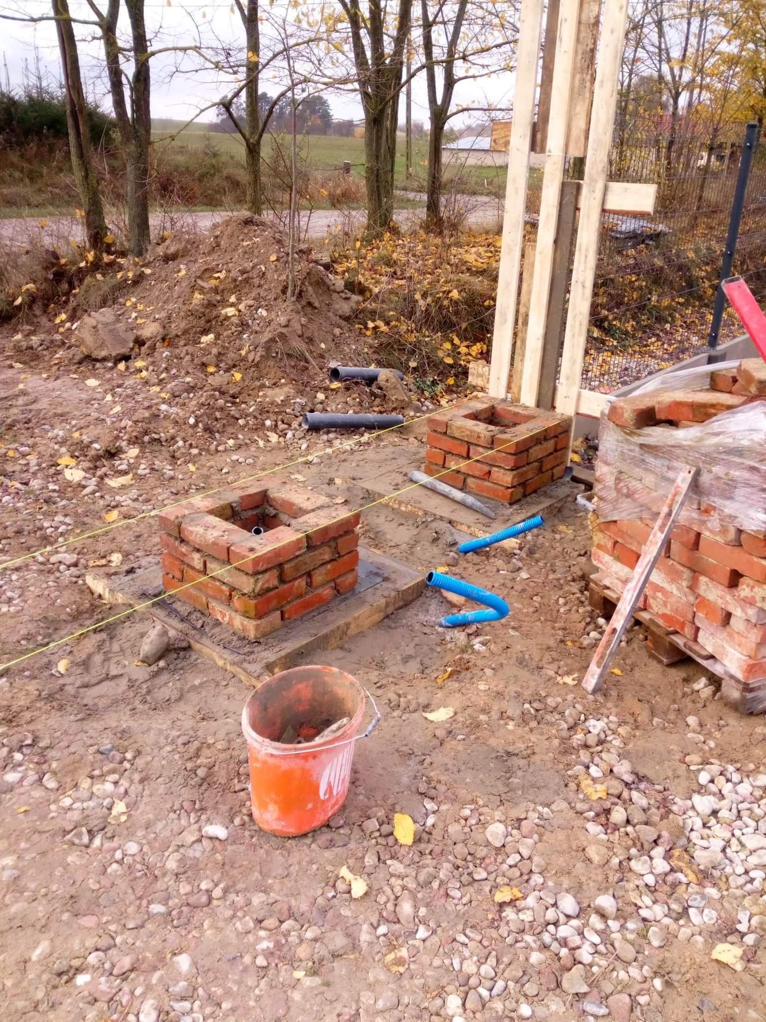 Dwa murowane, ceglane fundamenty pod słupy ogrodzeniowe na drewnianych podkładach, z widocznymi rurami i poziomnicą, w tle drewniane słupki i jesienne drzewa.