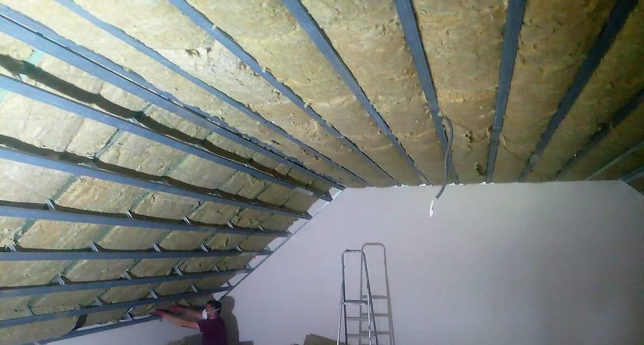 Montaż izolacji z wełny mineralnej na poddaszu, widoczne metalowe profile konstrukcyjne, drabina i pracownik w masce ochronnej.