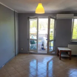 Pomieszczenie po malowaniu ścian na szaro, z widocznymi oknami balkonowymi i kaloryferem. Część mebli zabezpieczona folią malarską.