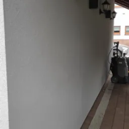 Świeżo pomalowana elewacja budynku z zamontowanymi lampami zewnętrznymi, widoczny fragment chodnika z kostki brukowej oraz myjka ciśnieniowa.