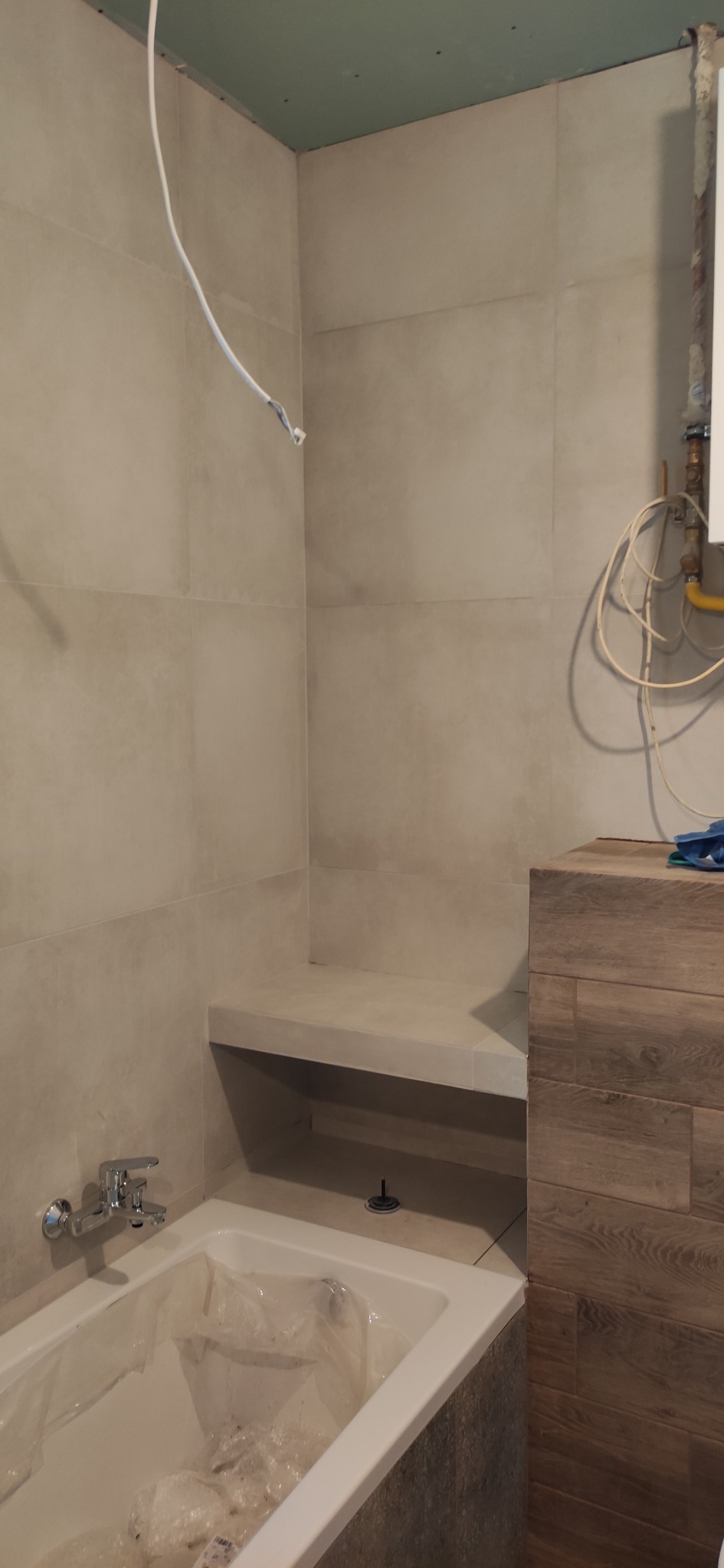 Łazienka w trakcie prac wykończeniowych: obudowana wanna z baterią, fragment siedziska z płytek, ściany wyłożone jasnymi płytkami, widoczne przewody elektryczne i instalacja grzewcza.