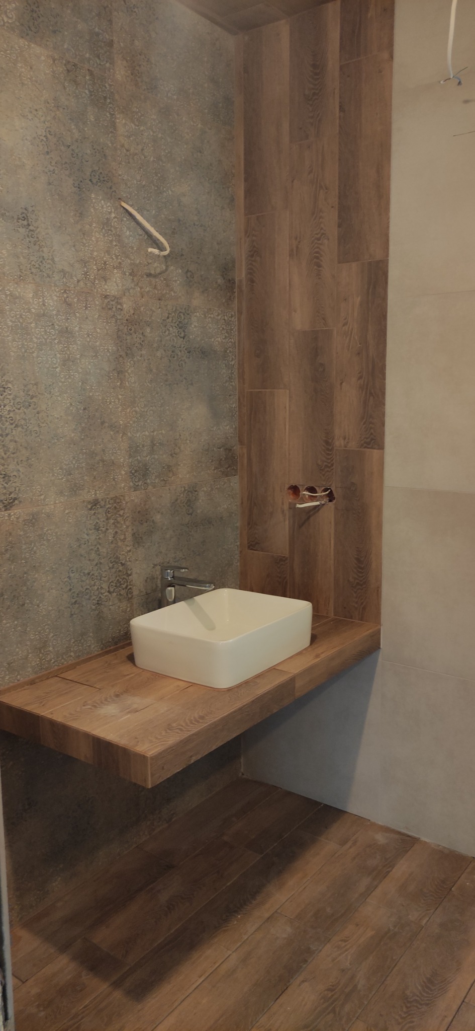 Nowoczesna, kwadratowa umywalka nablatowa z chromowaną baterią, zamontowana na drewnianym blacie w łazience z częściowo wykończonymi ścianami w odcieniach szarości i brązu.