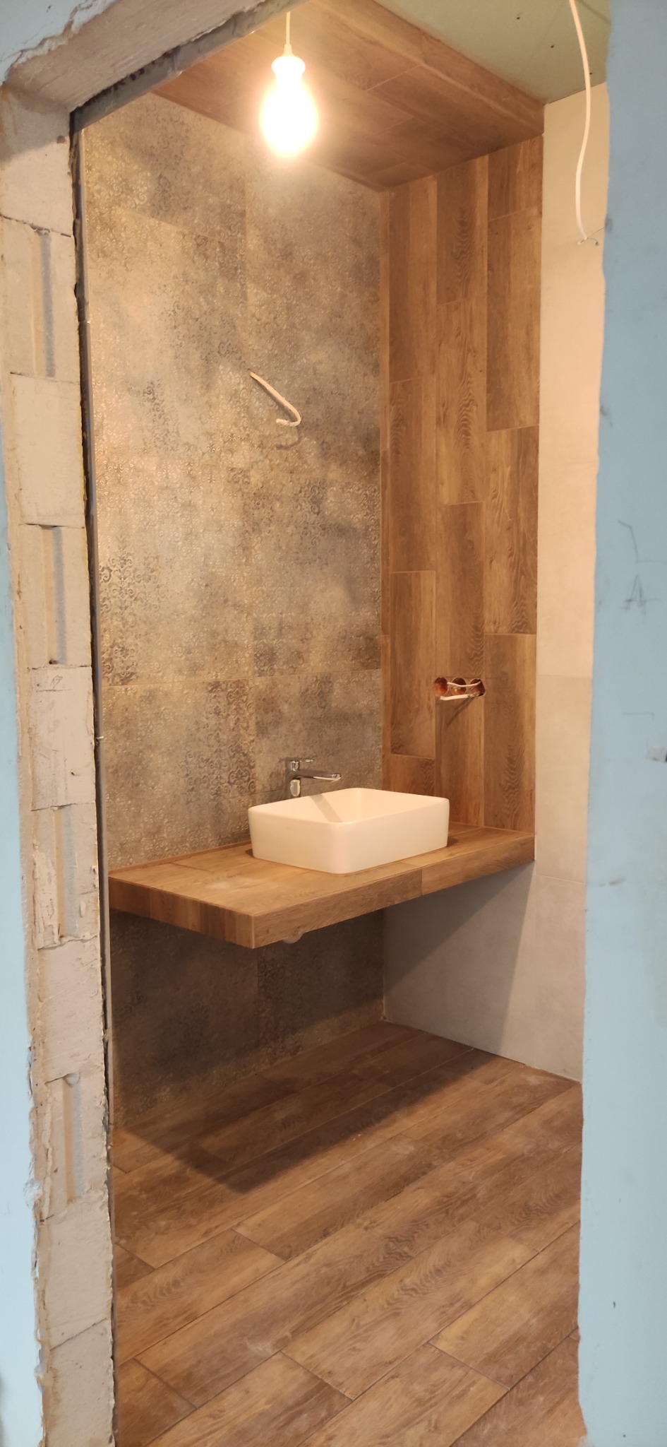 Łazienka w trakcie remontu: biała, prostokątna umywalka na drewnianym blacie, płytki imitujące drewno na podłodze i ścianie, widoczny hak oraz surowy stan wykończenia przy futrynie.