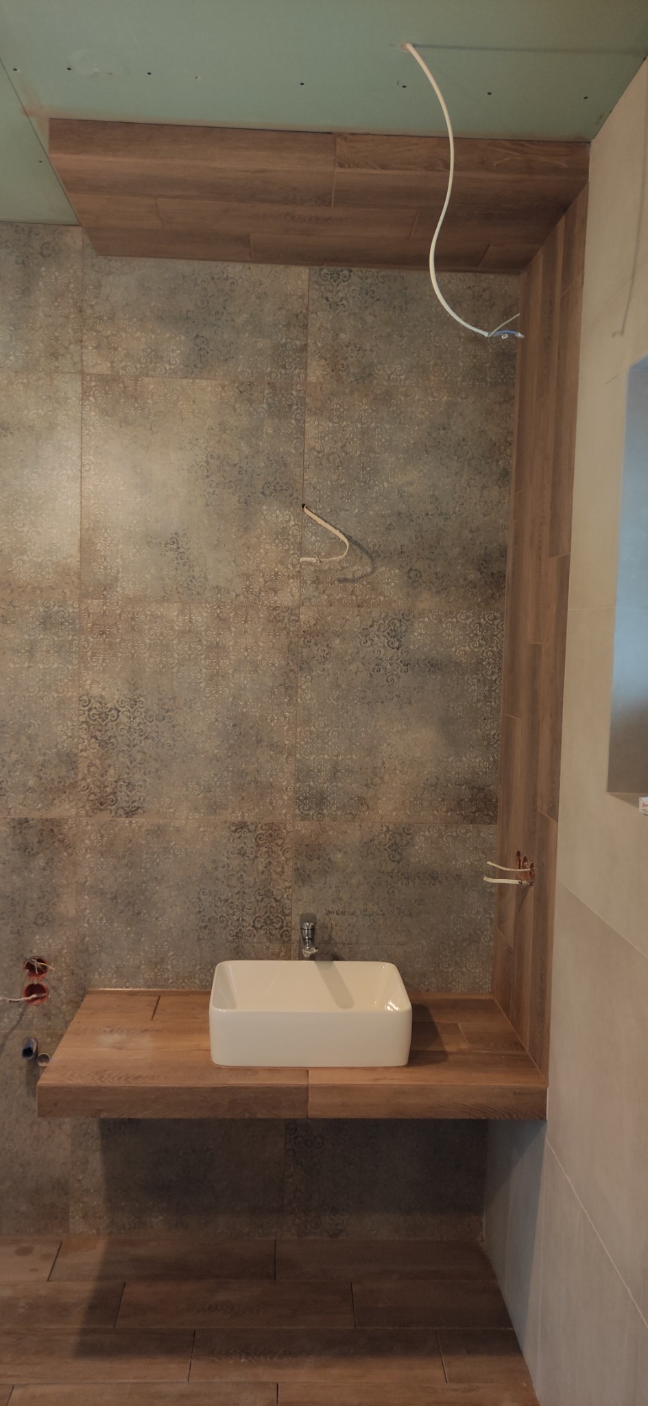 Łazienka w trakcie wykańczania: kwadratowa umywalka nablatowa, płytki ścienne z delikatnym wzorem, drewnopodobna zabudowa, wystające kable i rury.