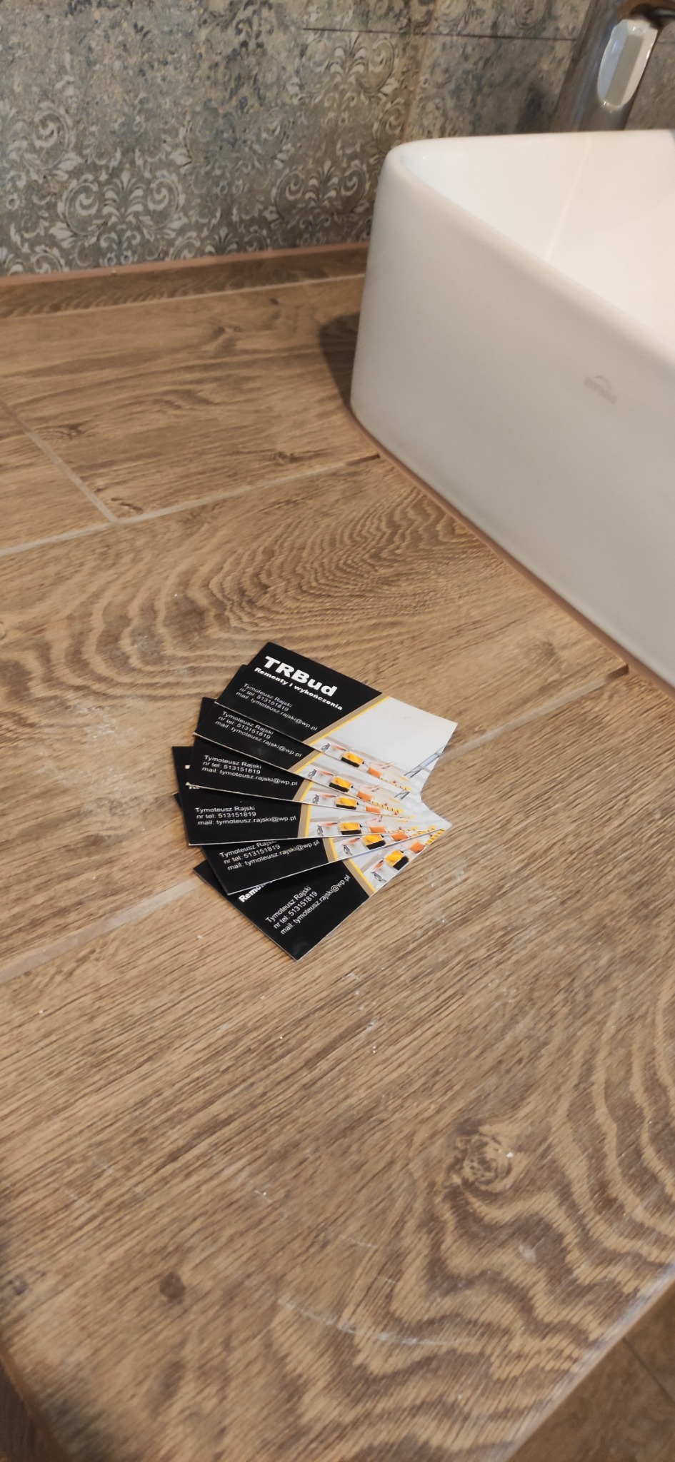 Stos wizytówek firmy remontowej TRBud leżący na drewnopodobnych panelach obok białej umywalki.