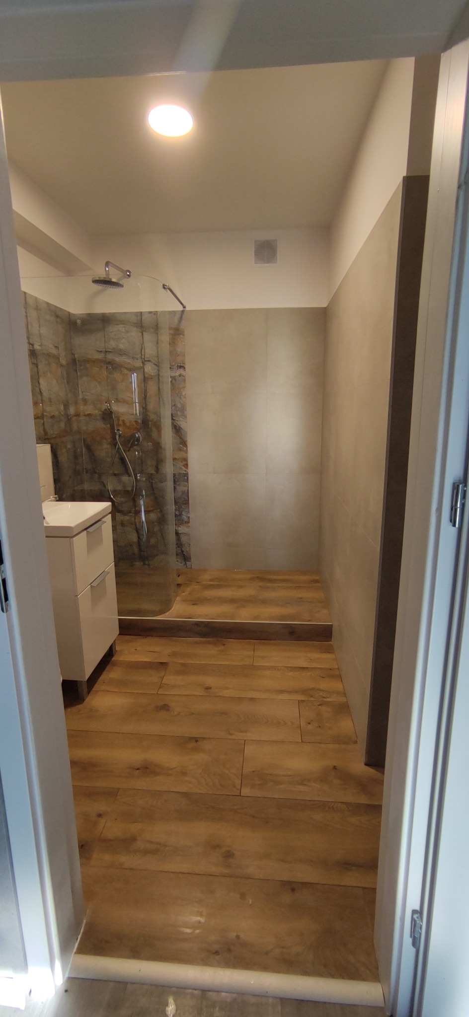 Nowoczesna łazienka z kabiną prysznicową typu walk-in, umywalką z szafką i drewnopodobną podłogą, widok zza uchylonych drzwi.
