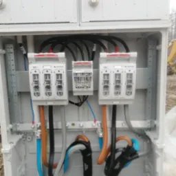 Otwarta skrzynka elektryczna z trzema bezpiecznikami RBK1 marki Apator, widoczne kolorowe przewody i ostrzeżenie o napięciu elektrycznym, na zewnątrz koparka.