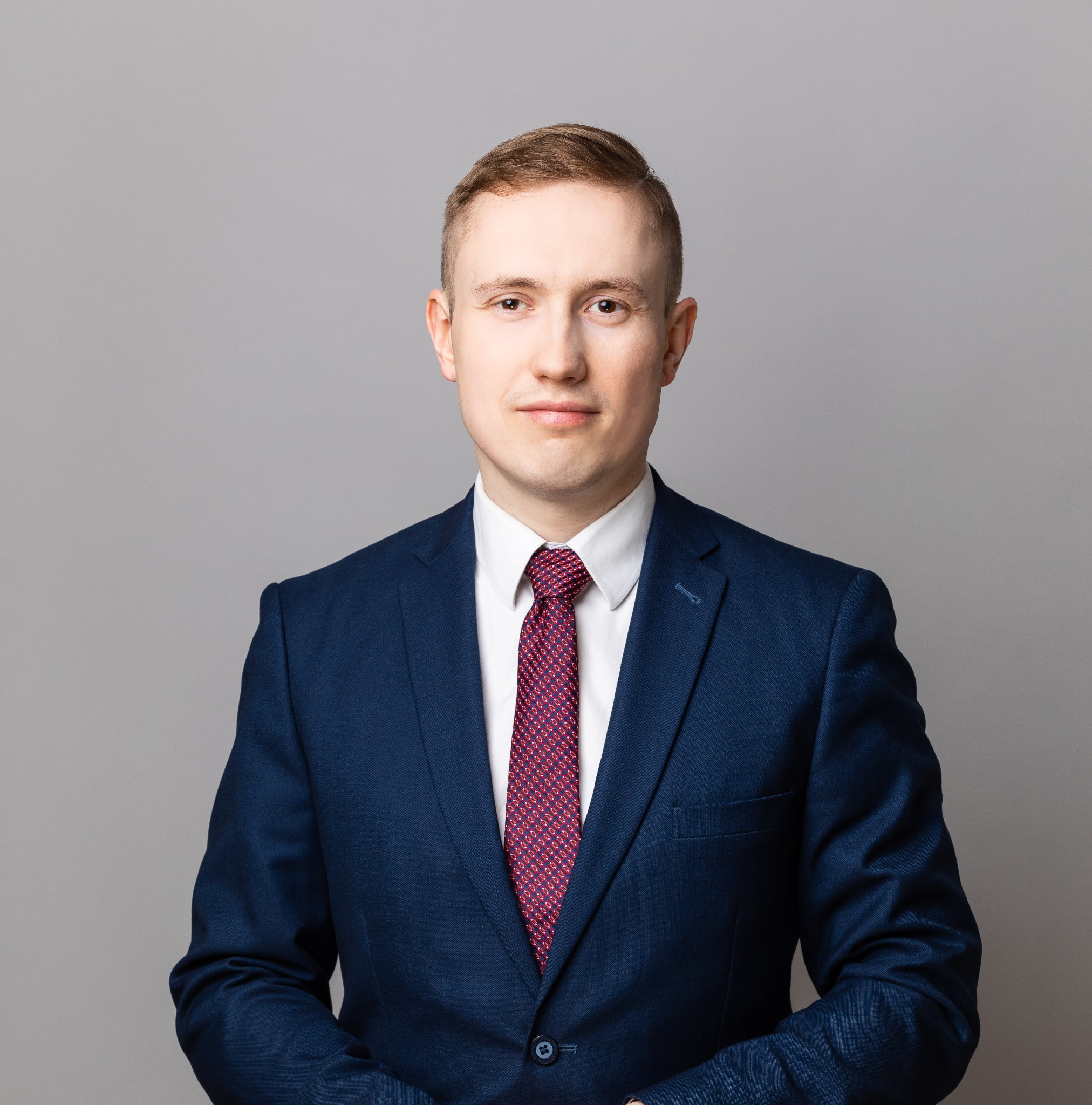 Portret mężczyzny w granatowym garniturze, białej koszuli i bordowym krawacie w geometryczne wzory, na szarym tle.