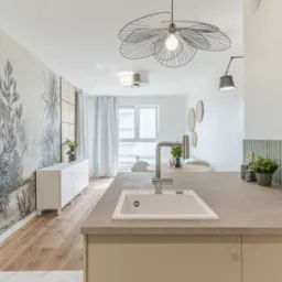 Jasne wnętrze: wzorzysta tapeta z motywem roślinnym, biała komoda, drewniana podłoga i designerska lampa nad stołem. W tle okrągłe lustra i zielona roślina.