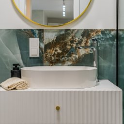 Molki Sp. z o.o. - Nowoczesna łazienka: owalna umywalka nablatowa na białej szafce z frezowanymi frontami, zielone płytki imitujące marmur i okrągłe lustro z złotą ramą.