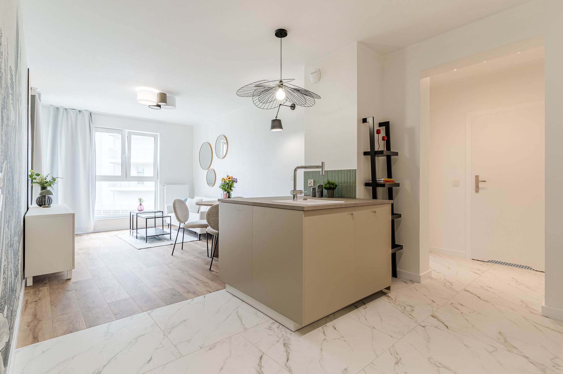 Jasne wnętrze po aranżacji w Gdańsku: marmurowe płytki, drewniana podłoga, wyspa kuchenna i salon z okrągłymi lustrami. Nowoczesne oświetlenie.