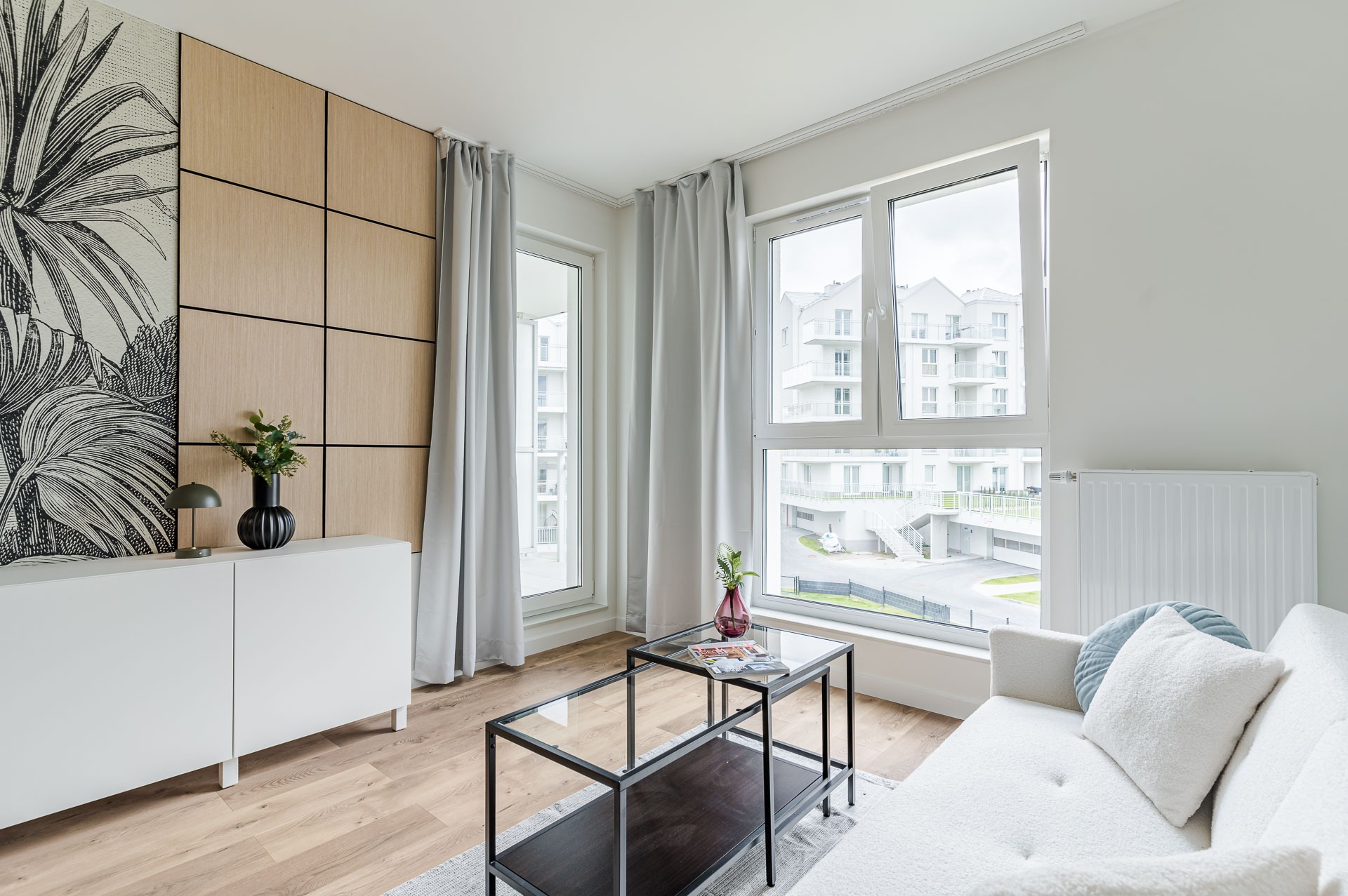 Jasny salon z drewnianą ścianą i wzorem liści, okna z widokiem na nowoczesne budynki, biała sofa i stolik kawowy. Minimalistyczny design i naturalne światło.