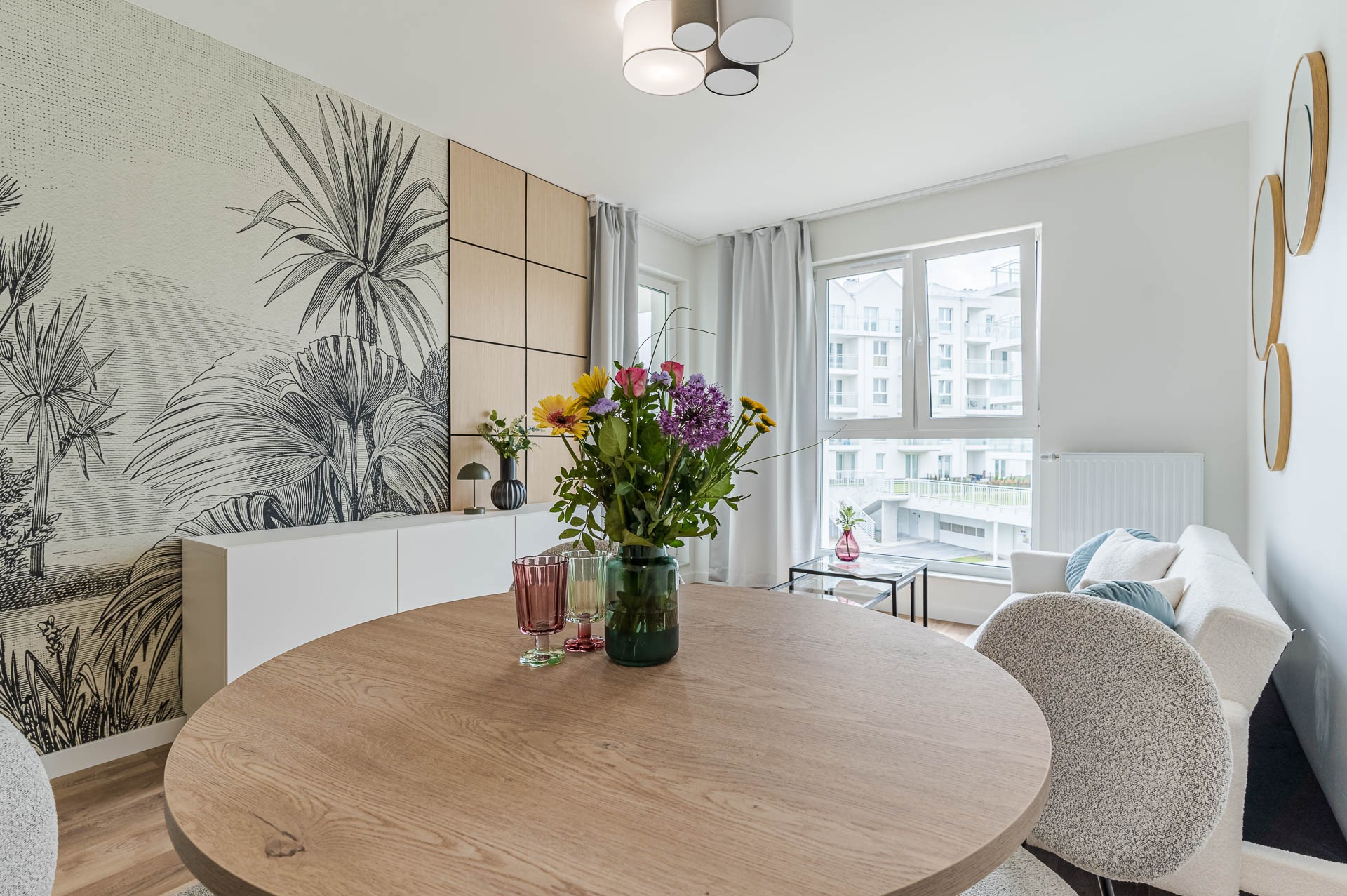 Jasne wnętrze jadalni z okrągłym stołem z drewna, kwiatami w wazonie i fototapetą z motywem roślinnym. Nowoczesne oświetlenie i minimalistyczny design.