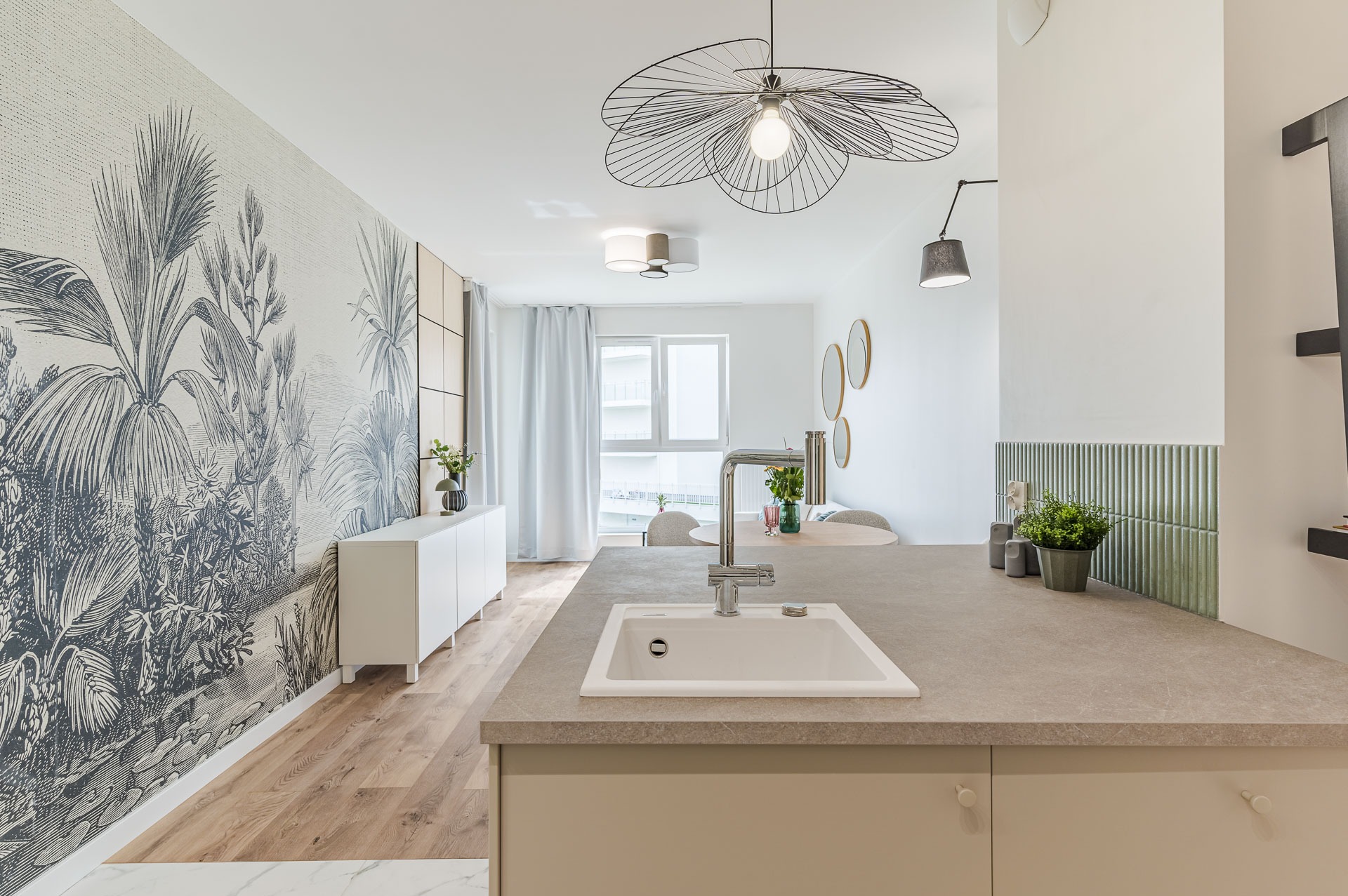 Jasne wnętrze: wzorzysta tapeta z motywem roślinnym, biała komoda, drewniana podłoga i designerska lampa nad stołem. W tle okrągłe lustra i zielona roślina.