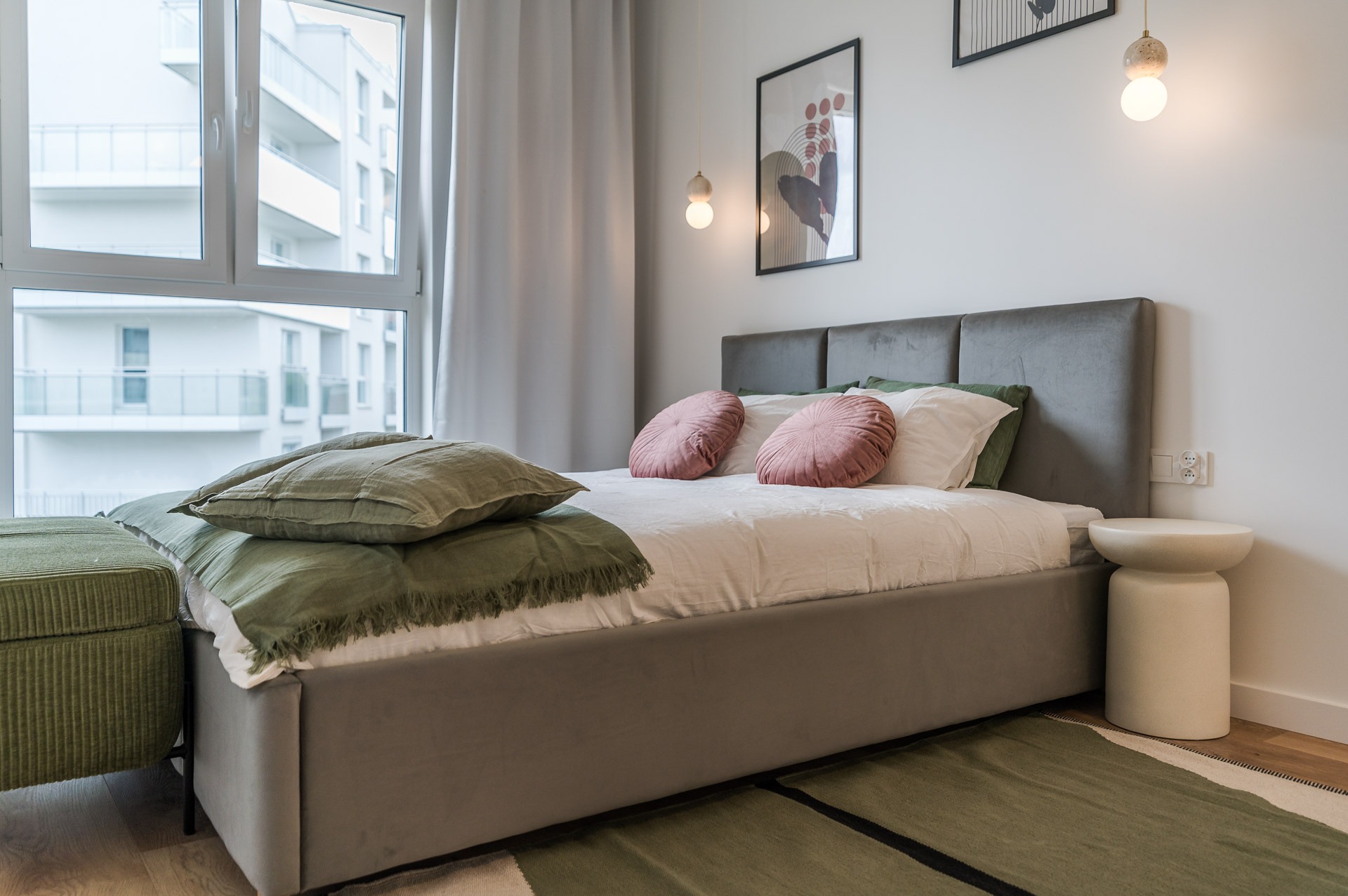 Nowoczesna sypialnia z łóżkiem kontynentalnym, zielonymi i różowymi poduszkami, minimalistyczną lampą i obrazami na ścianie. Widok na balkon zza okna.