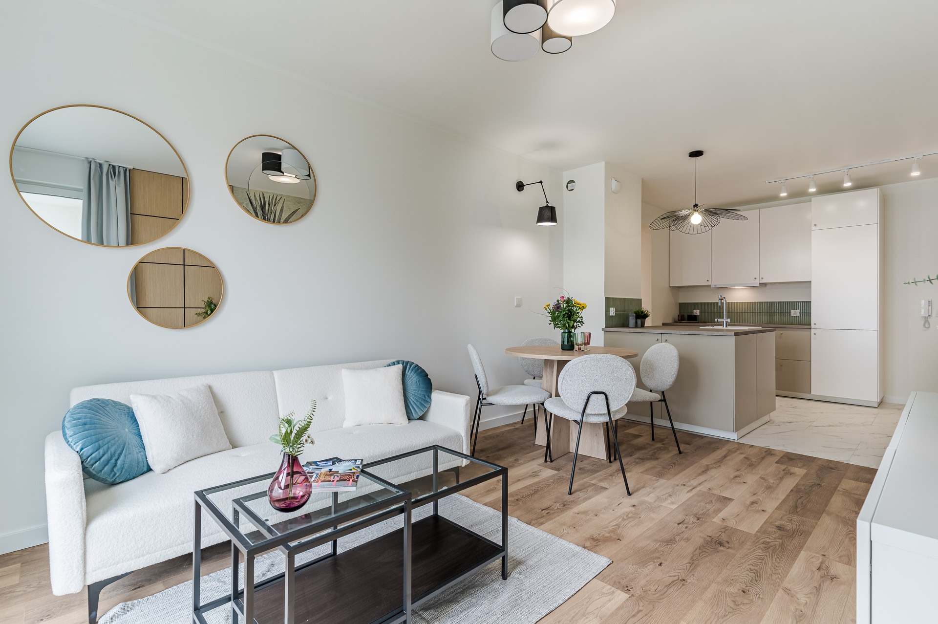 Jasne wnętrze: salon z sofą, stolikiem, okrągłym stołem i aneksem kuchennym. Na ścianie trzy okrągłe lustra. Parkiet na podłodze. Nowoczesny styl.