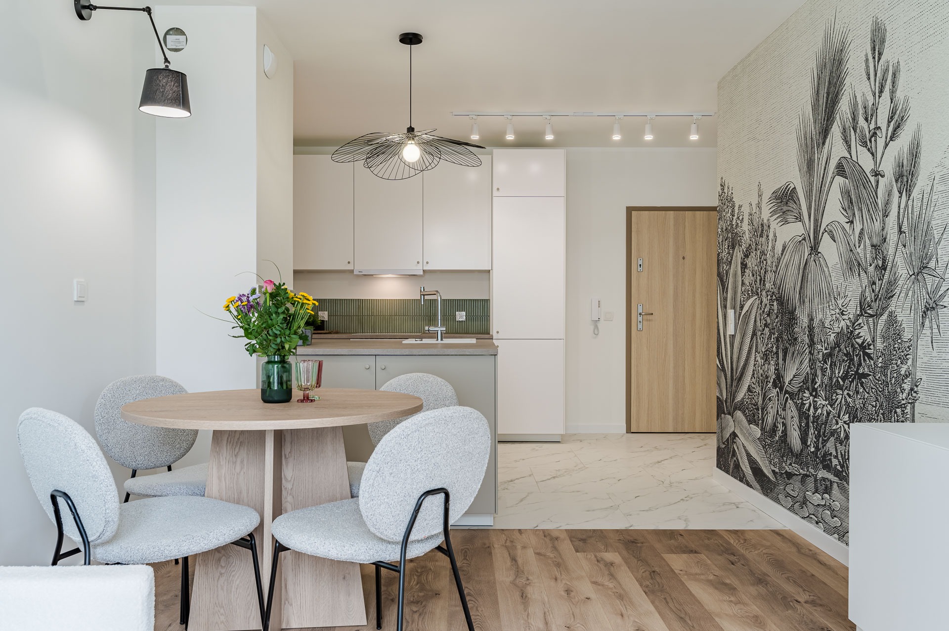 Jasne wnętrze: okrągły stół z bukietem, szare krzesła, kuchnia z zieloną mozaiką, fototapeta z motywem roślinnym. Nowoczesne oświetlenie.