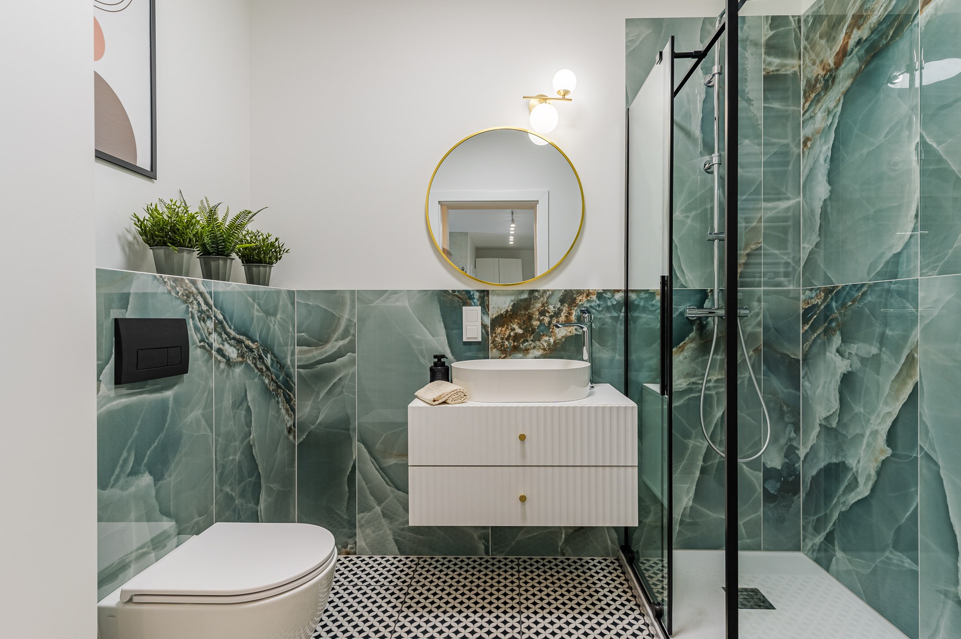 Nowoczesna łazienka z turkusowymi płytkami imitującymi marmur, okrągłym lustrem w złotej ramie i kabiną prysznicową typu walk-in. Czarno-biała podłoga w geometryczny wzór.