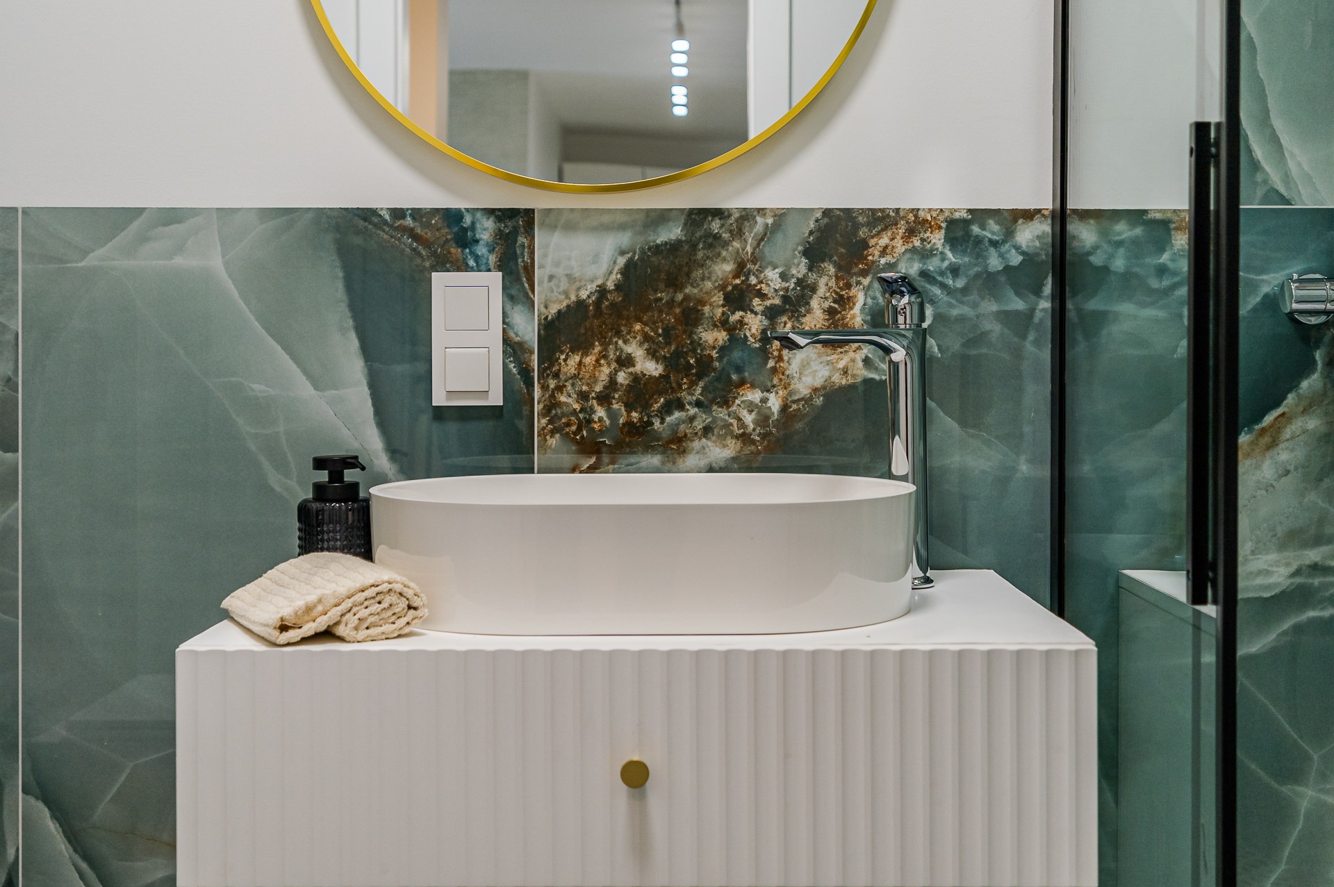 Nowoczesna łazienka: owalna umywalka nablatowa na białej szafce z frezowanymi frontami, zielone płytki imitujące marmur i okrągłe lustro z złotą ramą.