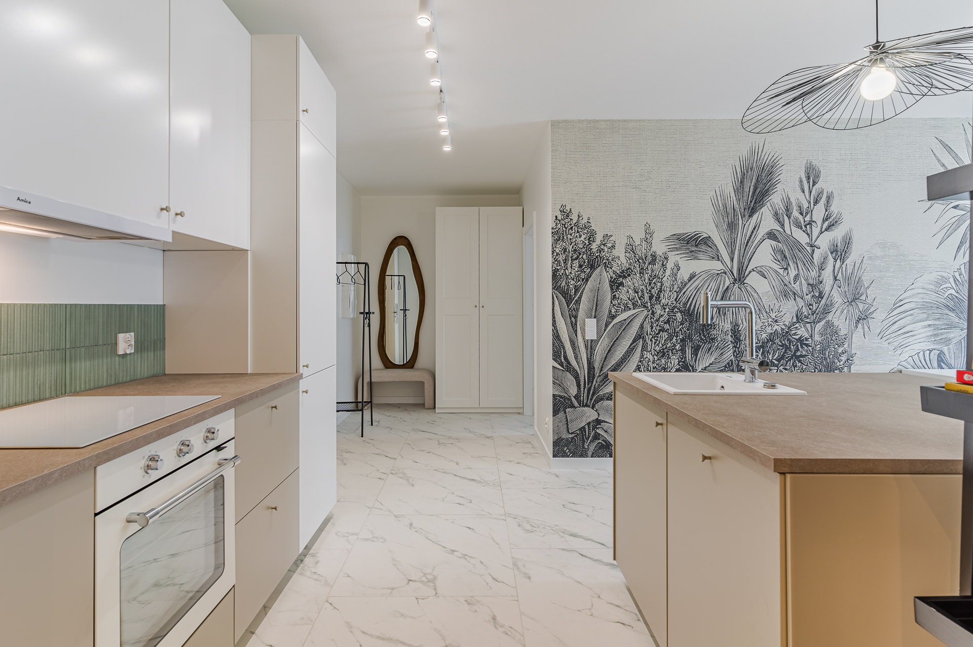 Jasna kuchnia z wyspą i tapetą w motywy roślinne. Minimalistyczne meble, marmurowa podłoga, oświetlenie punktowe i dekoracyjna lampa nad wyspą.