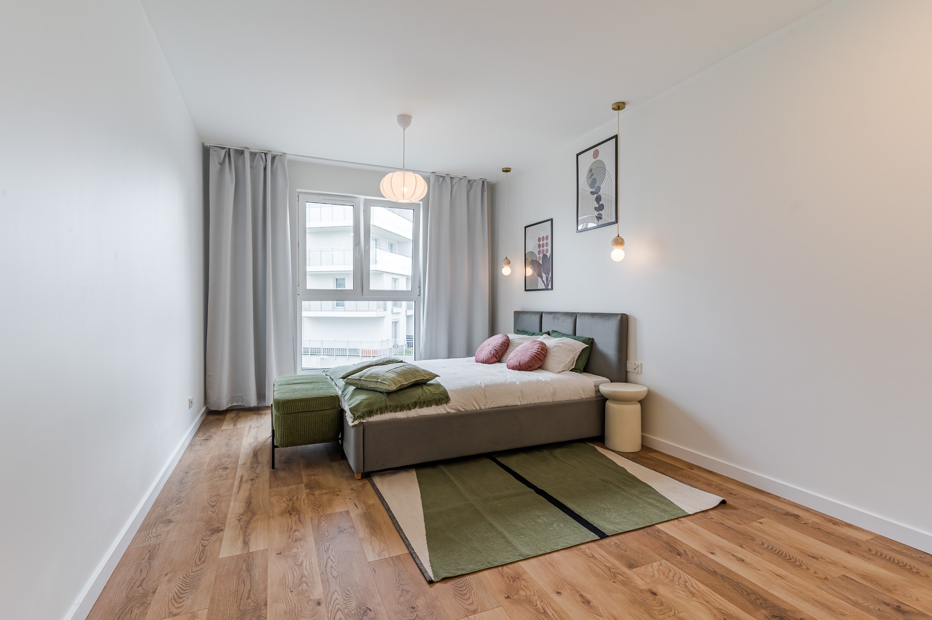 Nowoczesna sypialnia z łóżkiem, zielonym dywanem i pufą. Drewniana podłoga, minimalistyczne oświetlenie i grafiki na ścianie. Stonowana kolorystyka, jasne wnętrze.