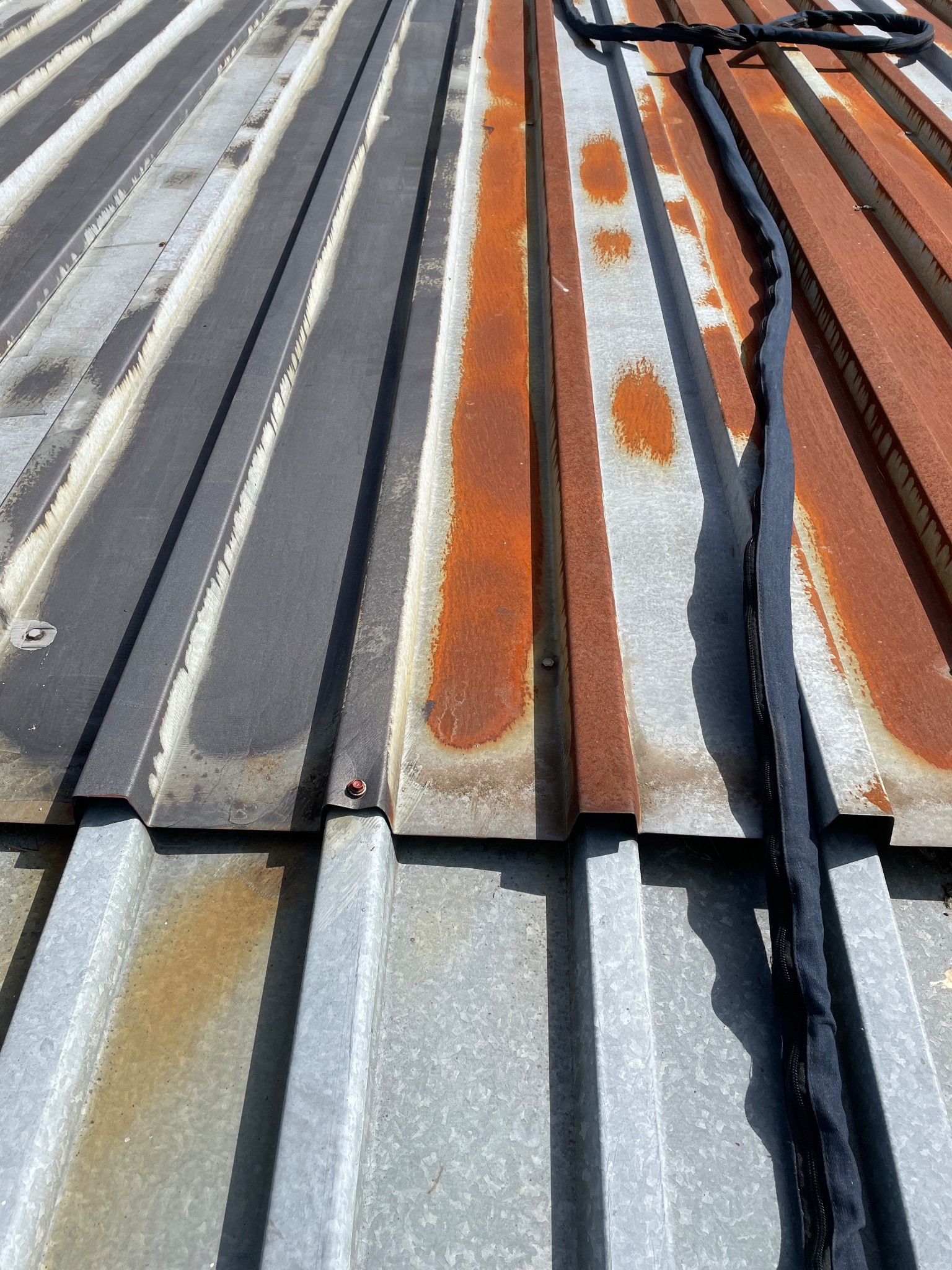 Metalowy dach z widocznymi śladami rdzy i czyszczenia, z czarnym przewodem leżącym na powierzchni, ukazujący kontrast między obszarami oczyszczonymi i zardzewiałymi.