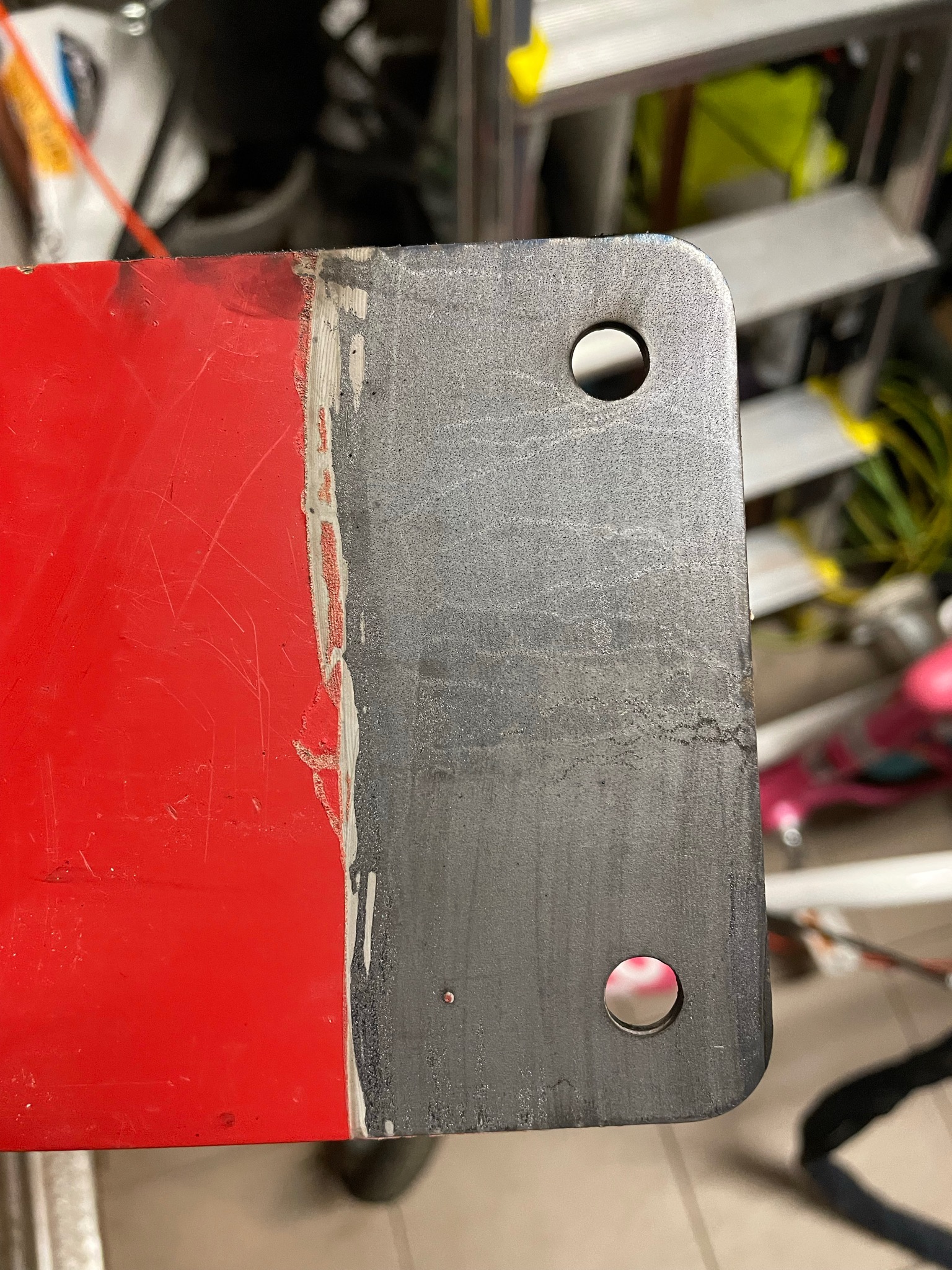 Metalowy element po piaskowaniu i częściowym usunięciu czerwonej farby, widoczne dwa otwory montażowe i ślady korozji na powierzchni.