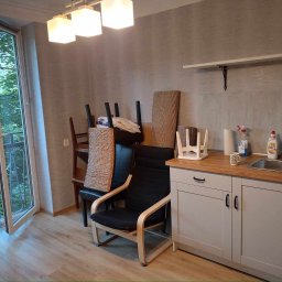 USŁUGI BUDOWLANE PAWEŁ LAUTER - Wnętrze pokoju z otwartym balkonem, widoczna zabudowa kuchenna z drewnianym blatem, krzesła ustawione obok, na podłodze panele imitujące drewno.